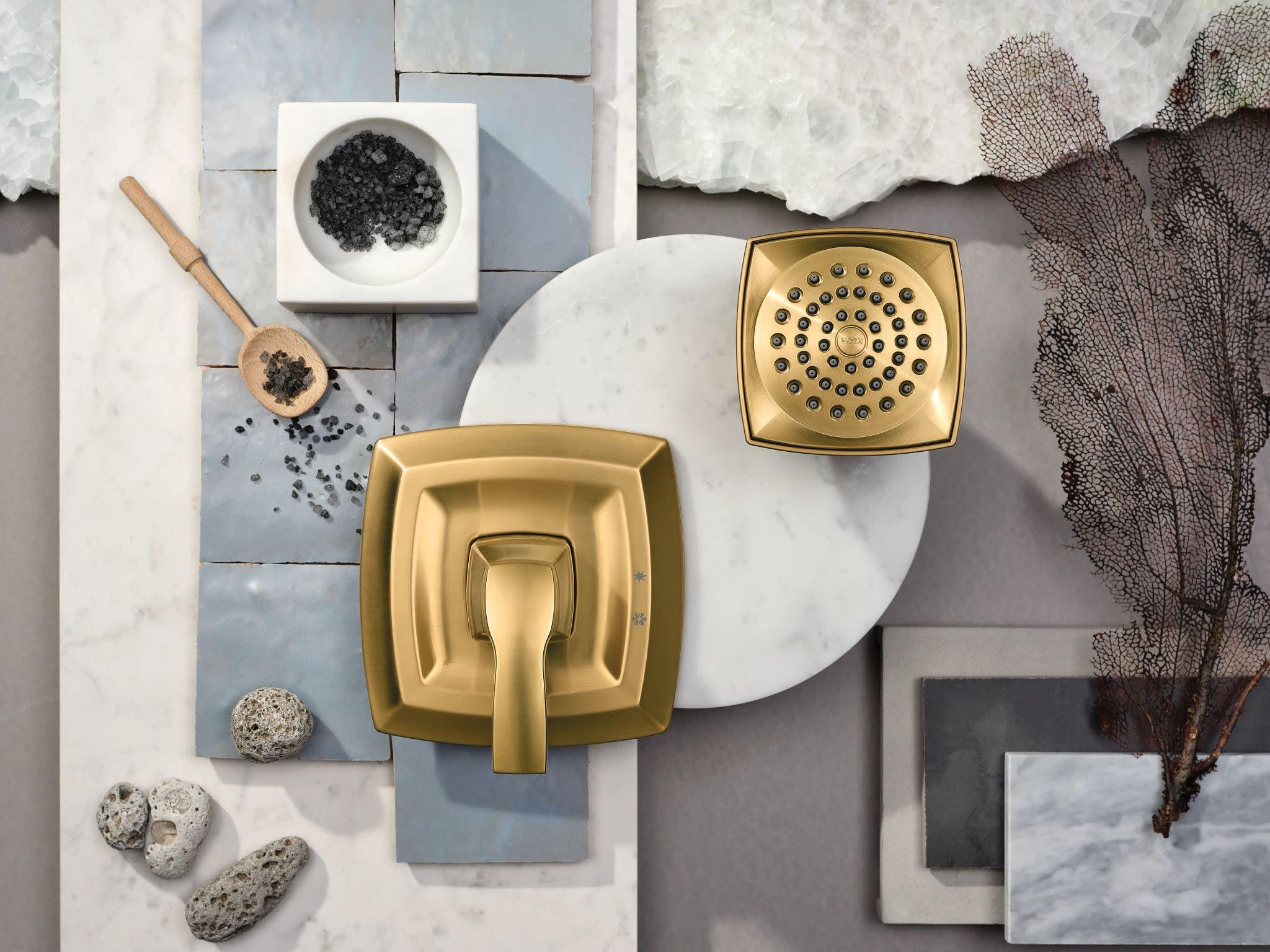Gold-finished shower valve trim kit with square handle and matching showerhead, displayed with bathroom accessories.