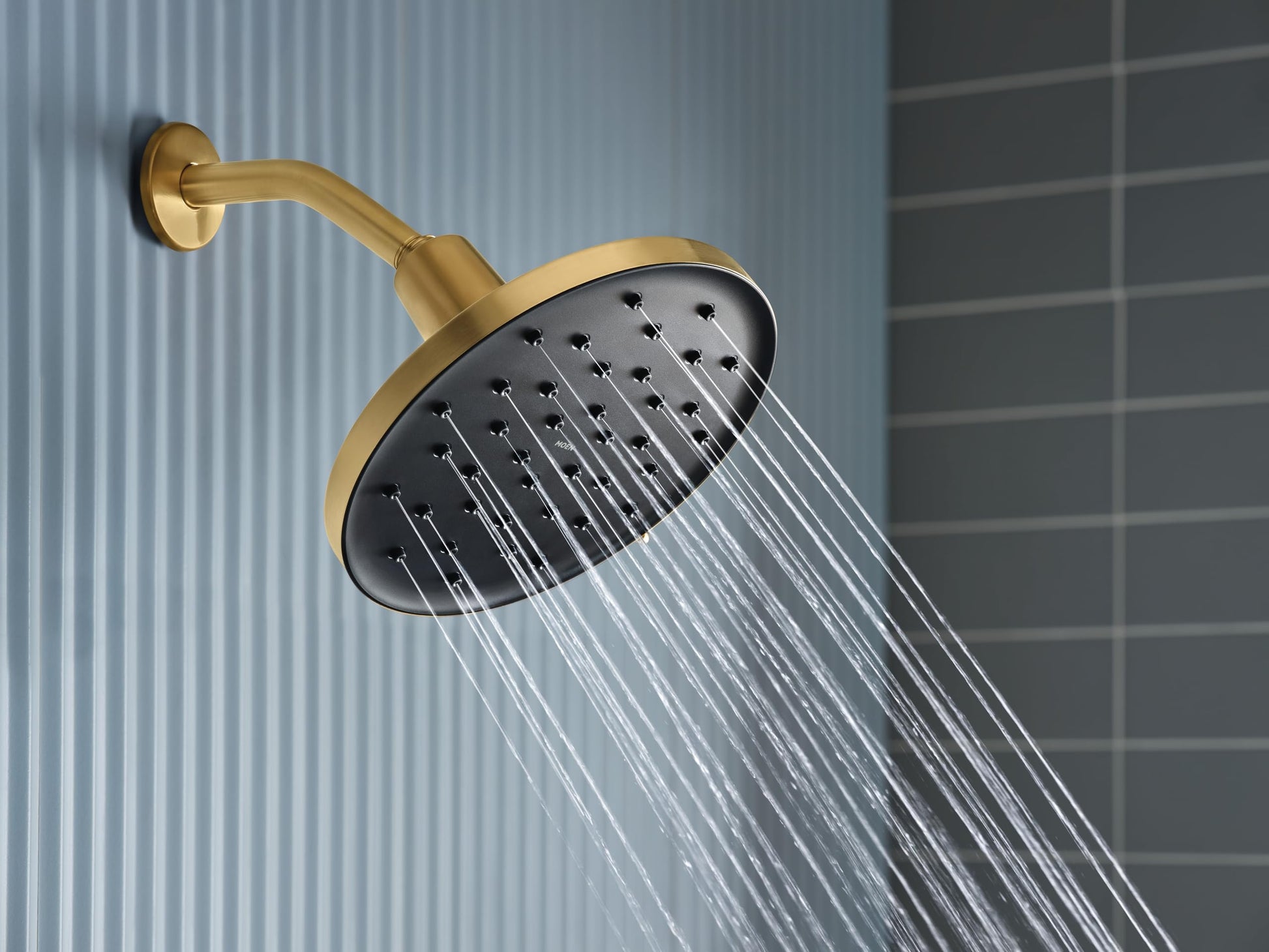 Brushed gold rainfall showerhead with black faceplate and curved arm mounted on tile wall with water streaming downward.