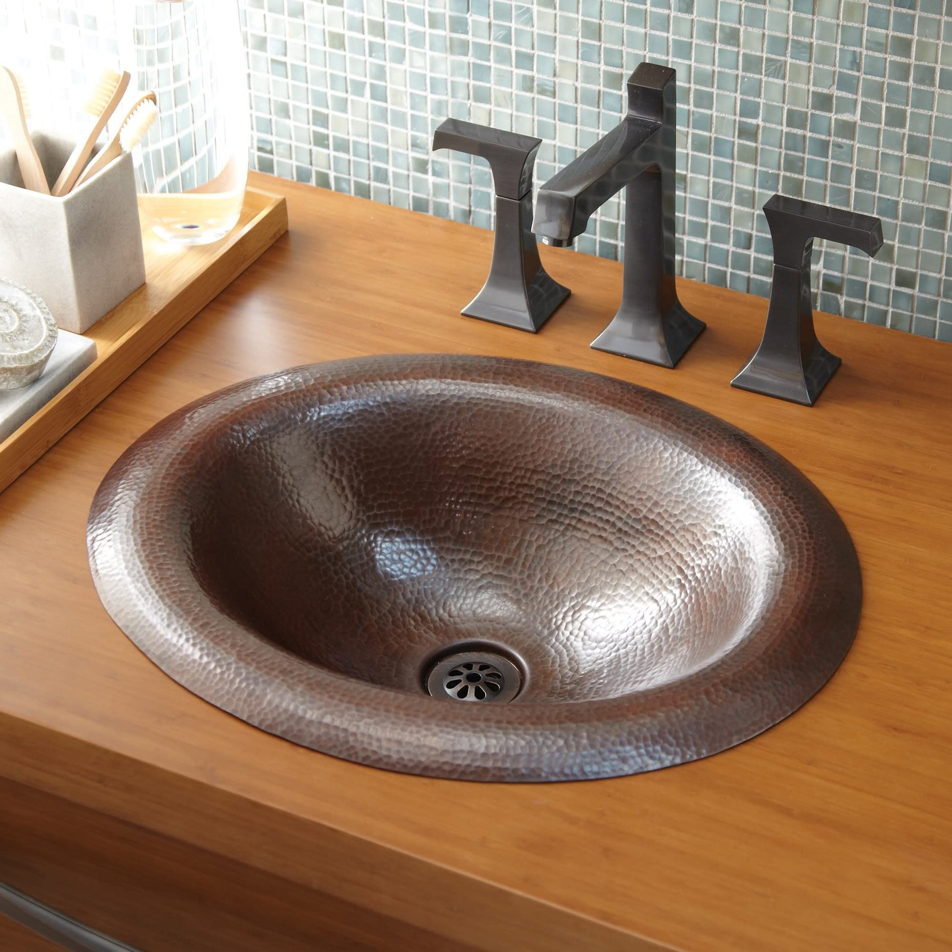 Copper-finish oval bathroom sink with hammered texture and three black faucet handles on wooden vanity.