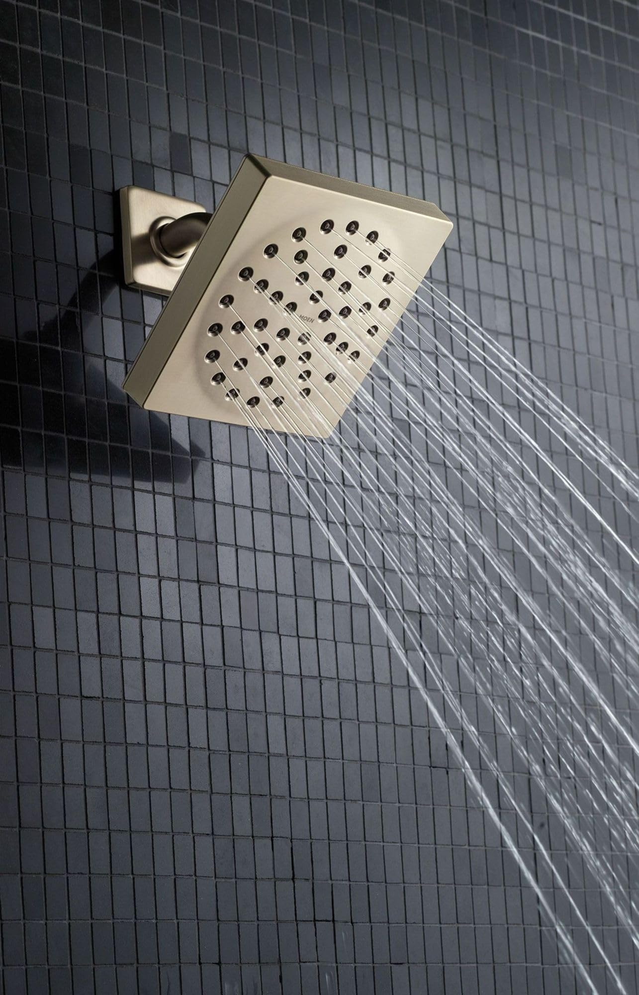Chrome square showerhead with multiple spray holes mounted at an angle on tile wall with water streaming downward.