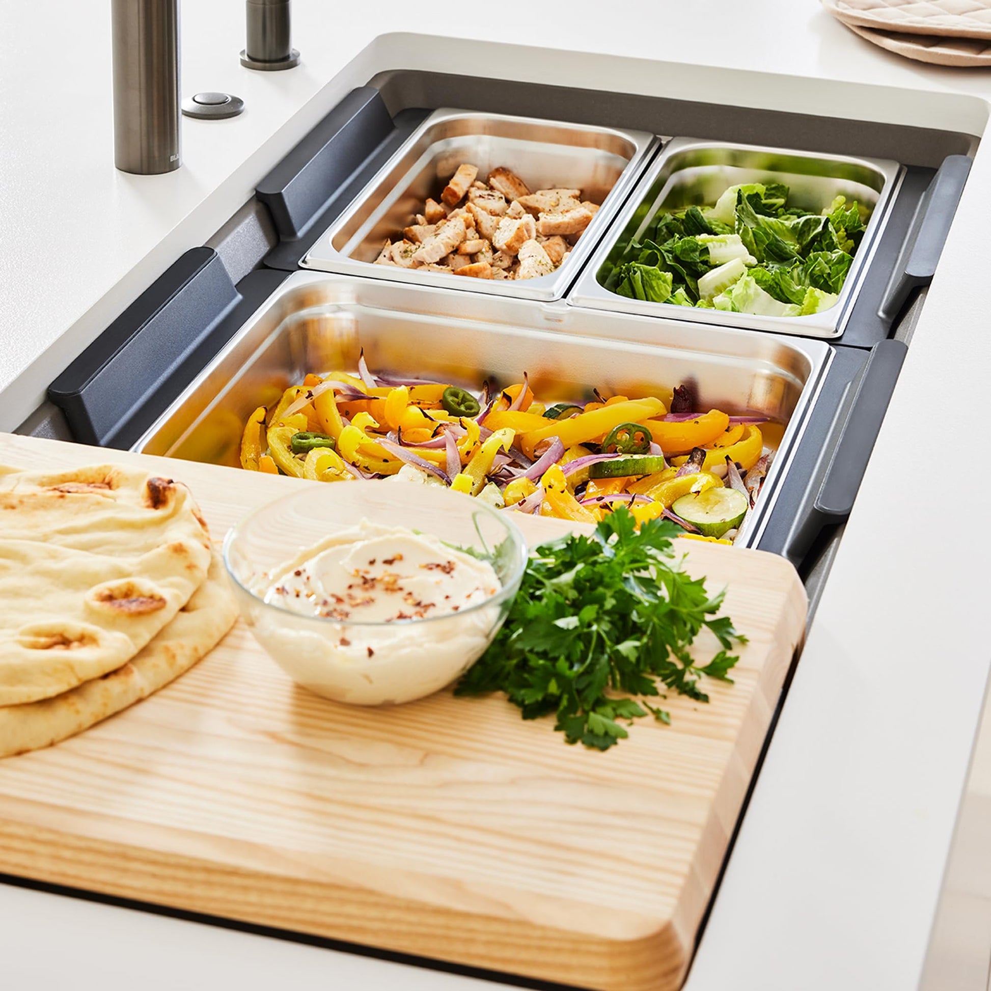 Stainless steel sink tray with three food containers holding prepared ingredients and wooden cutting board in sink.