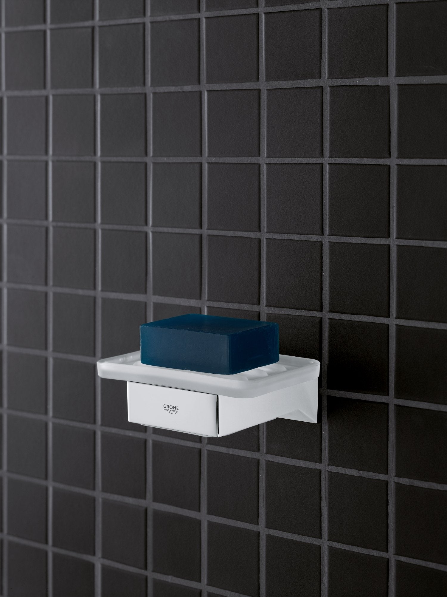 Chrome soap dish with white rectangular base and blue soap bar on dark tiled bathroom wall.