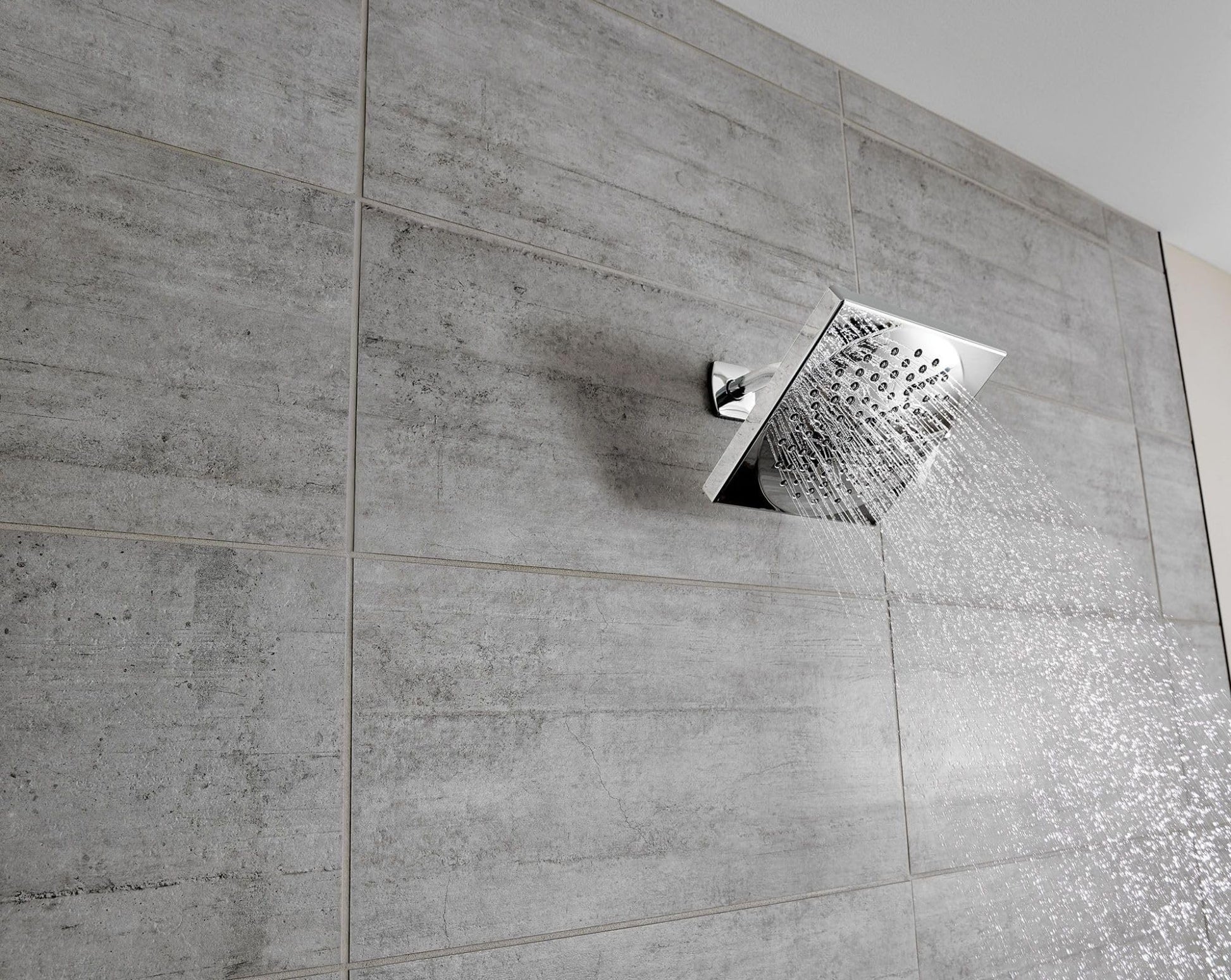 Brushed nickel square rainshower showerhead mounted on gray tile wall with water spray visible.