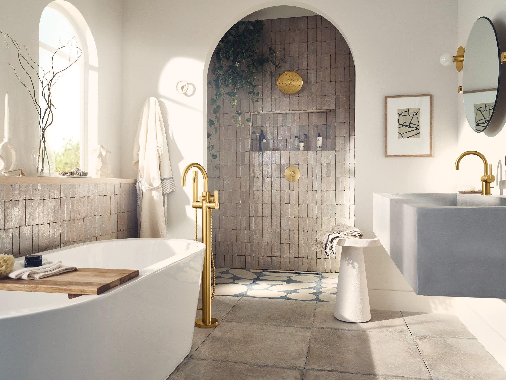 Brushed gold freestanding tub filler with single handle and hand shower, installed beside white soaking tub in bathroom.