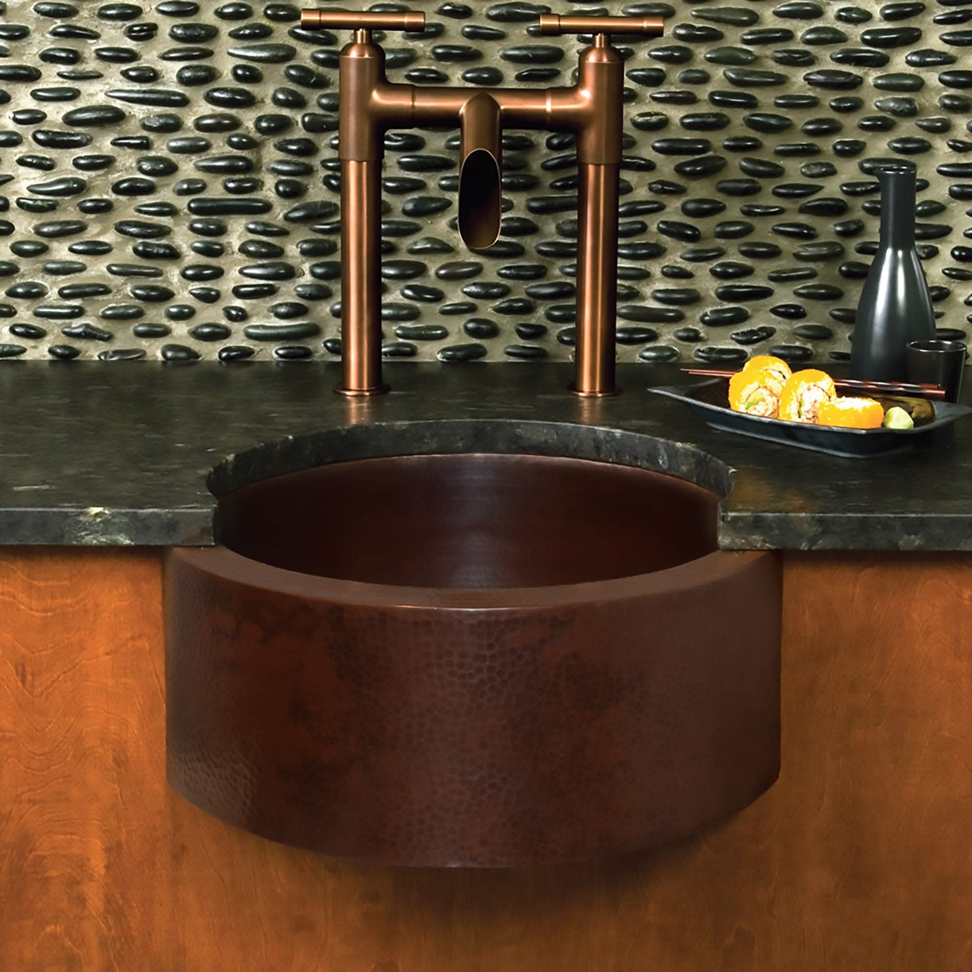 Hammered copper undermount sink with cylindrical bowl and copper bridge faucet installed in countertop.