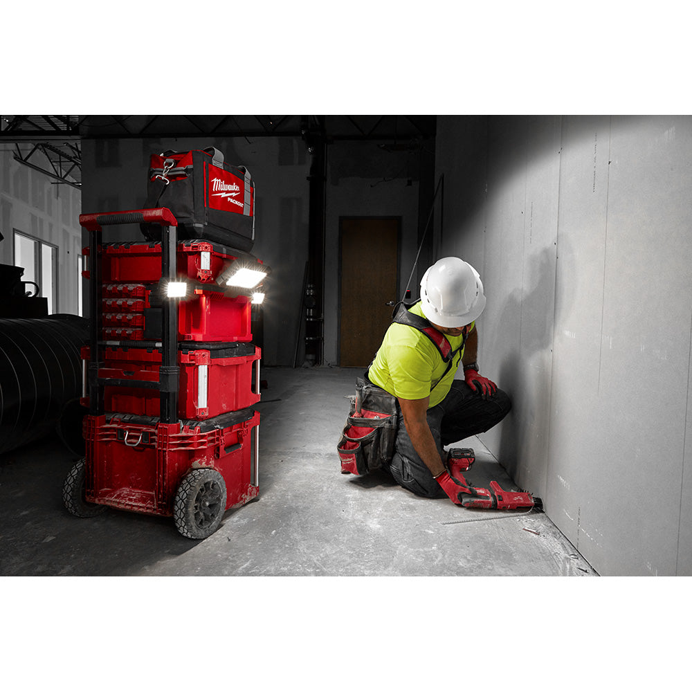 Worker in safety gear wearing red and black tool bag while crouching against wall in industrial setting.