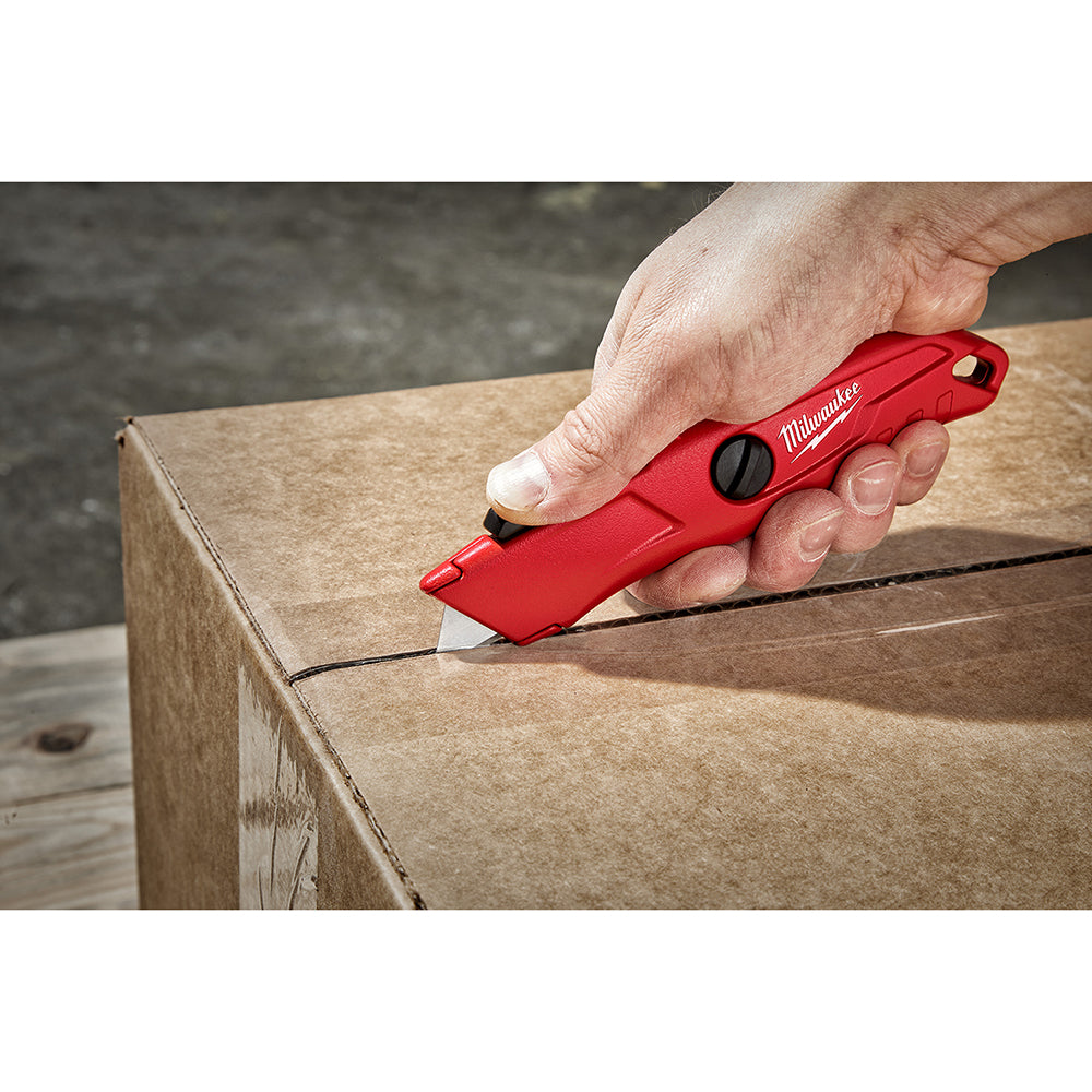 Red Milwaukee self-retracting utility knife with black button mechanism, held in hand cutting cardboard box.