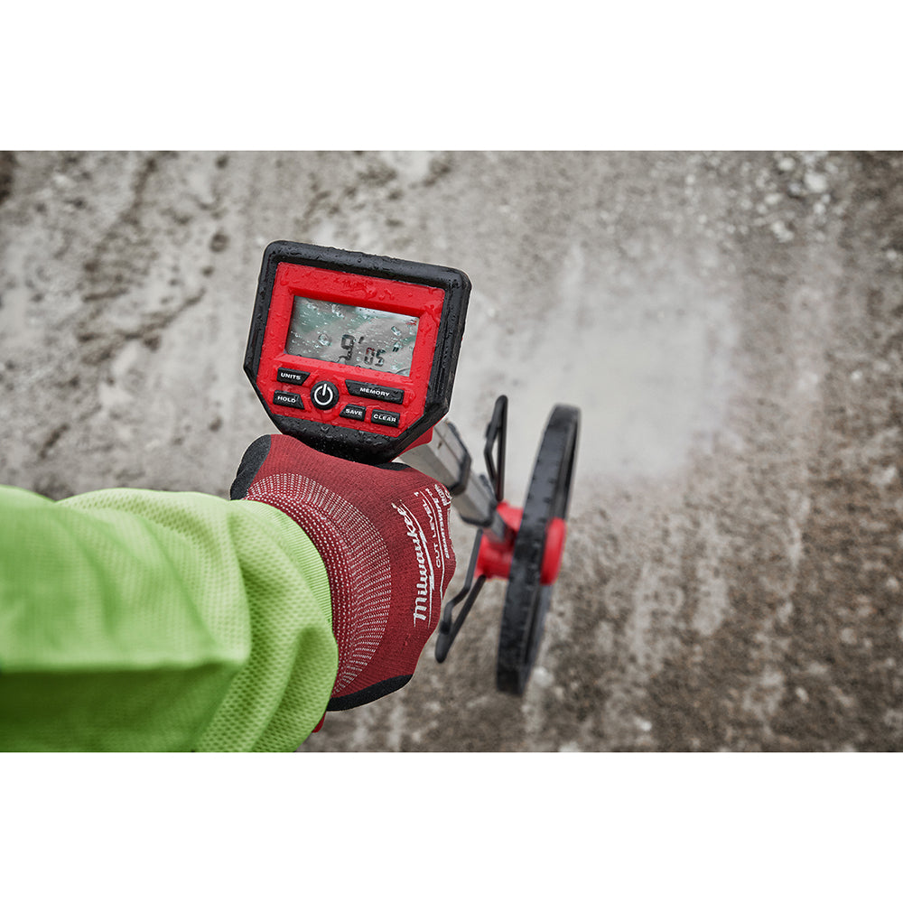 Red and black digital measuring wheel with LCD display held in hand above concrete surface.