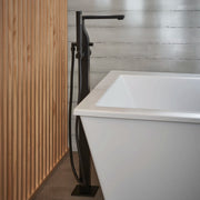Matte black floor-mounted bathtub filler with square spout, hand shower, and hose next to white soaking tub.