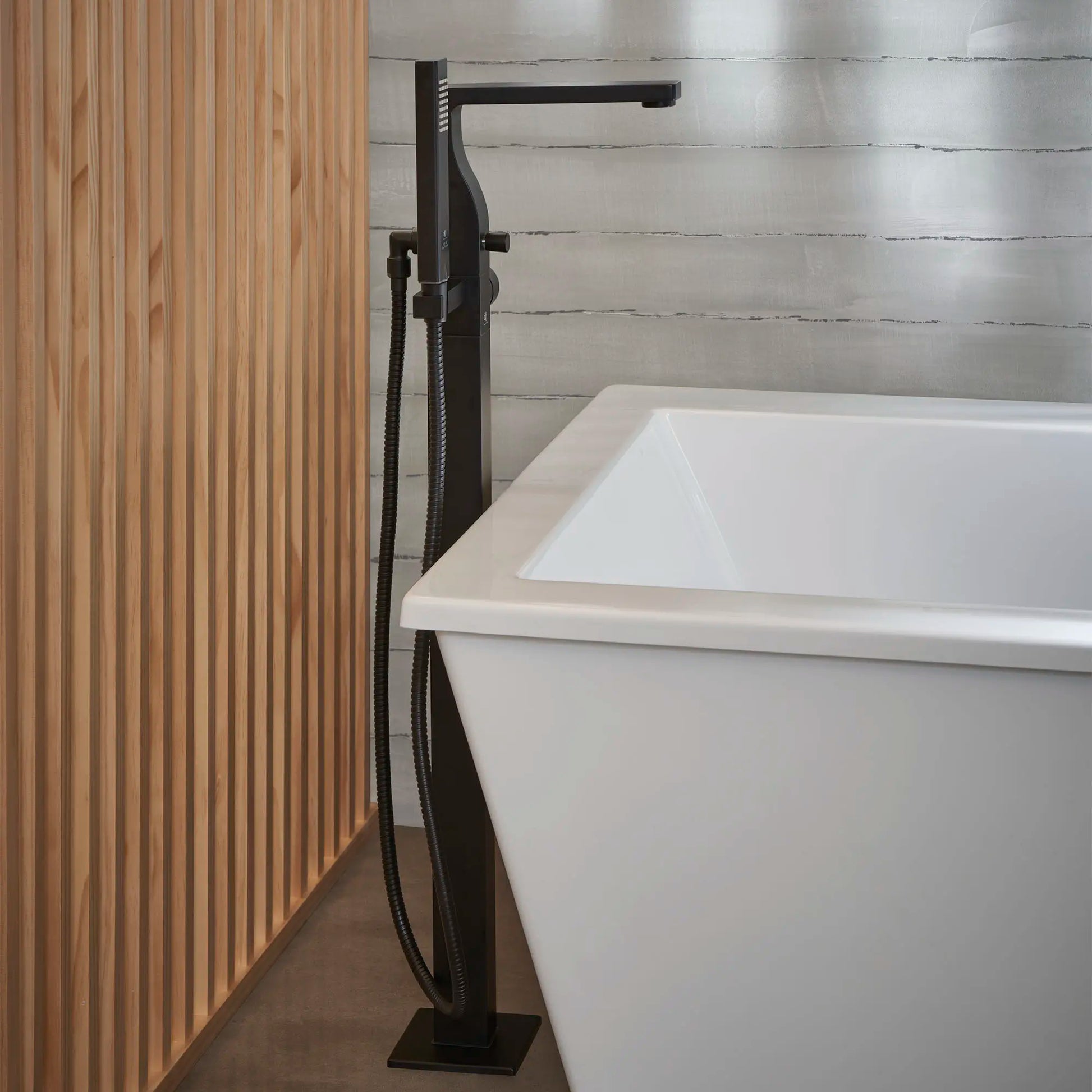 Matte black floor-mounted bathtub filler with square spout, hand shower, and hose next to white soaking tub.