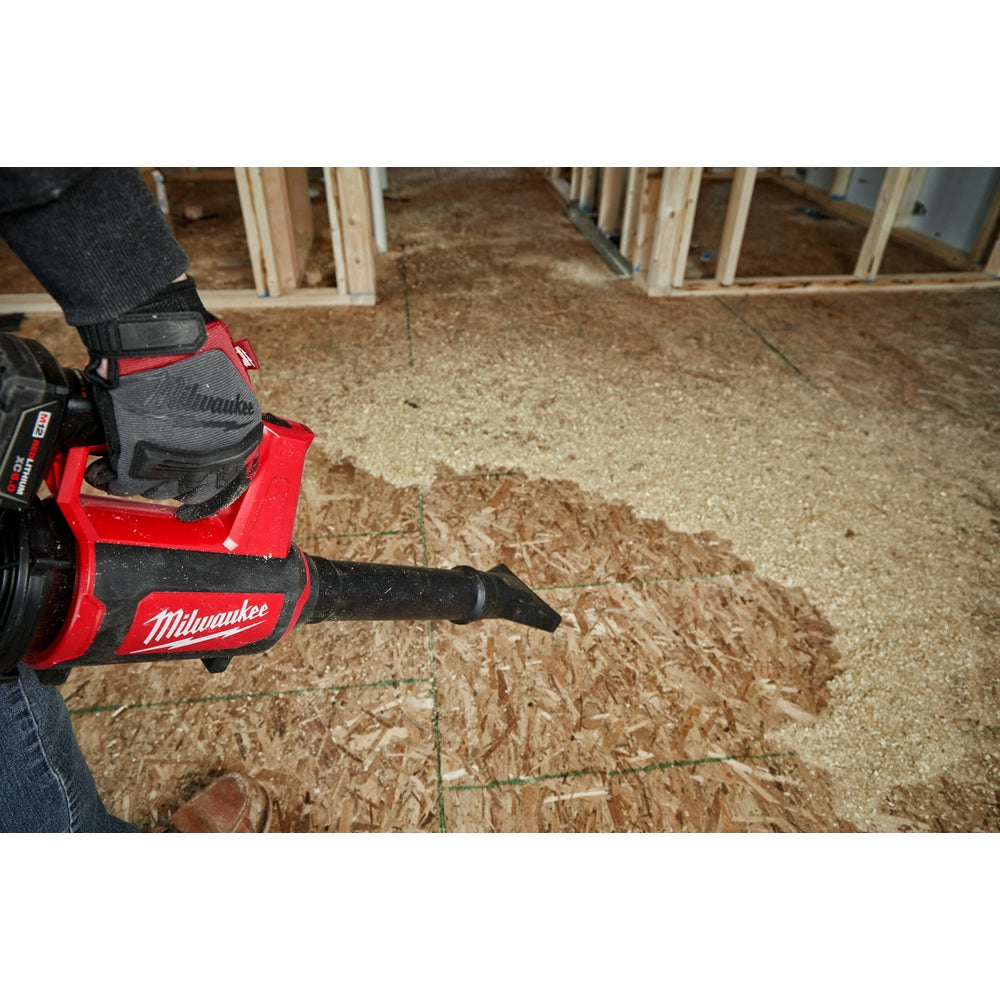Red and black Milwaukee M12 compact blower being held by a person wearing work gloves, blowing sawdust on a concrete floor.