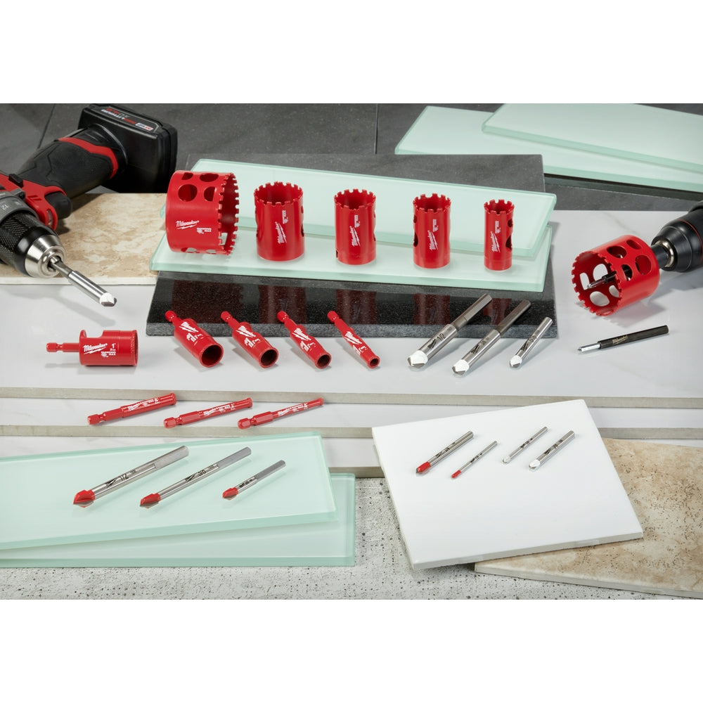 Silver carbide drill bit with red tip displayed among Milwaukee drilling accessories and hole saws on work surface.