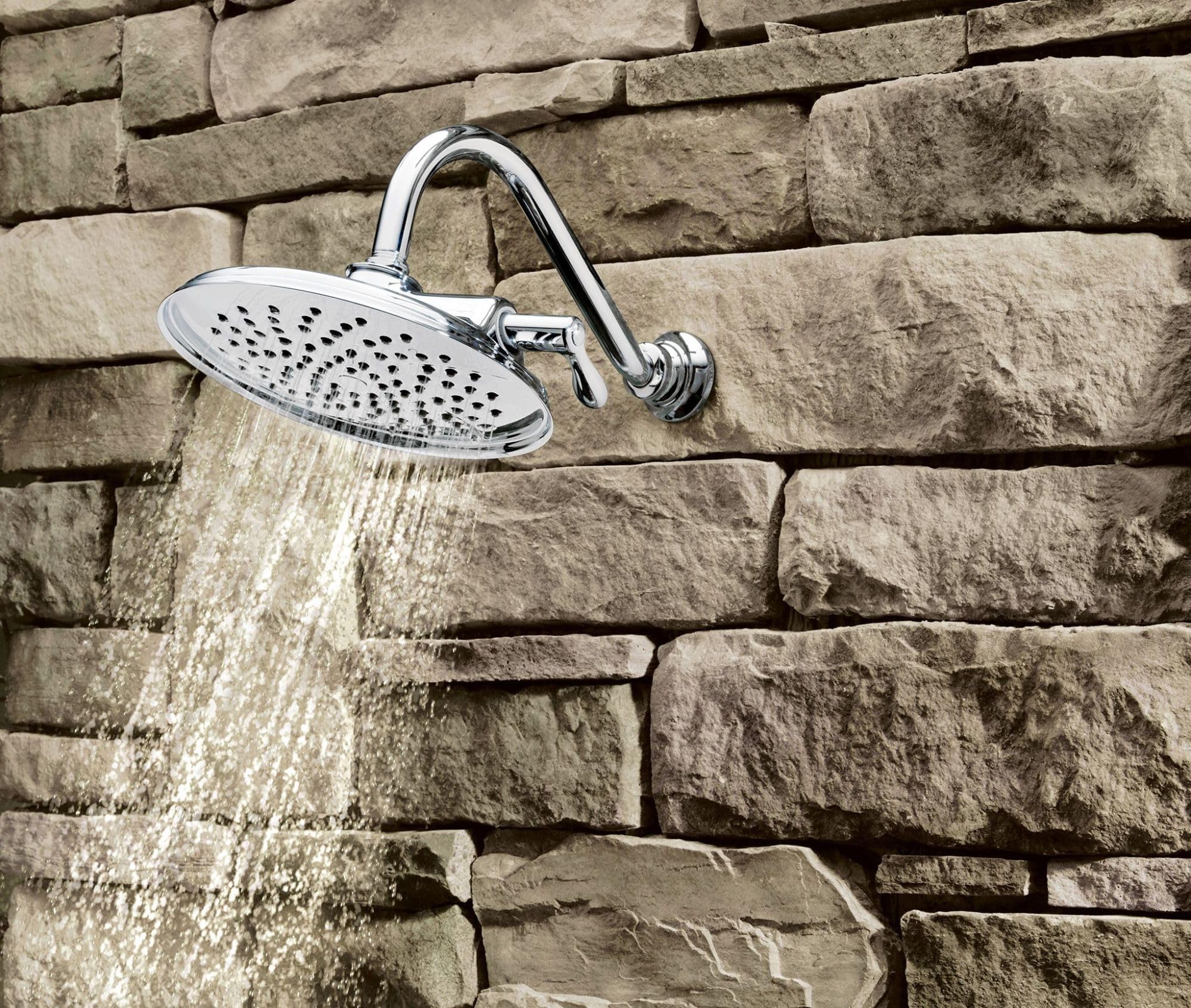 Chrome showerhead with oval face and perforated spray pattern mounted on curved arm against stone wall.