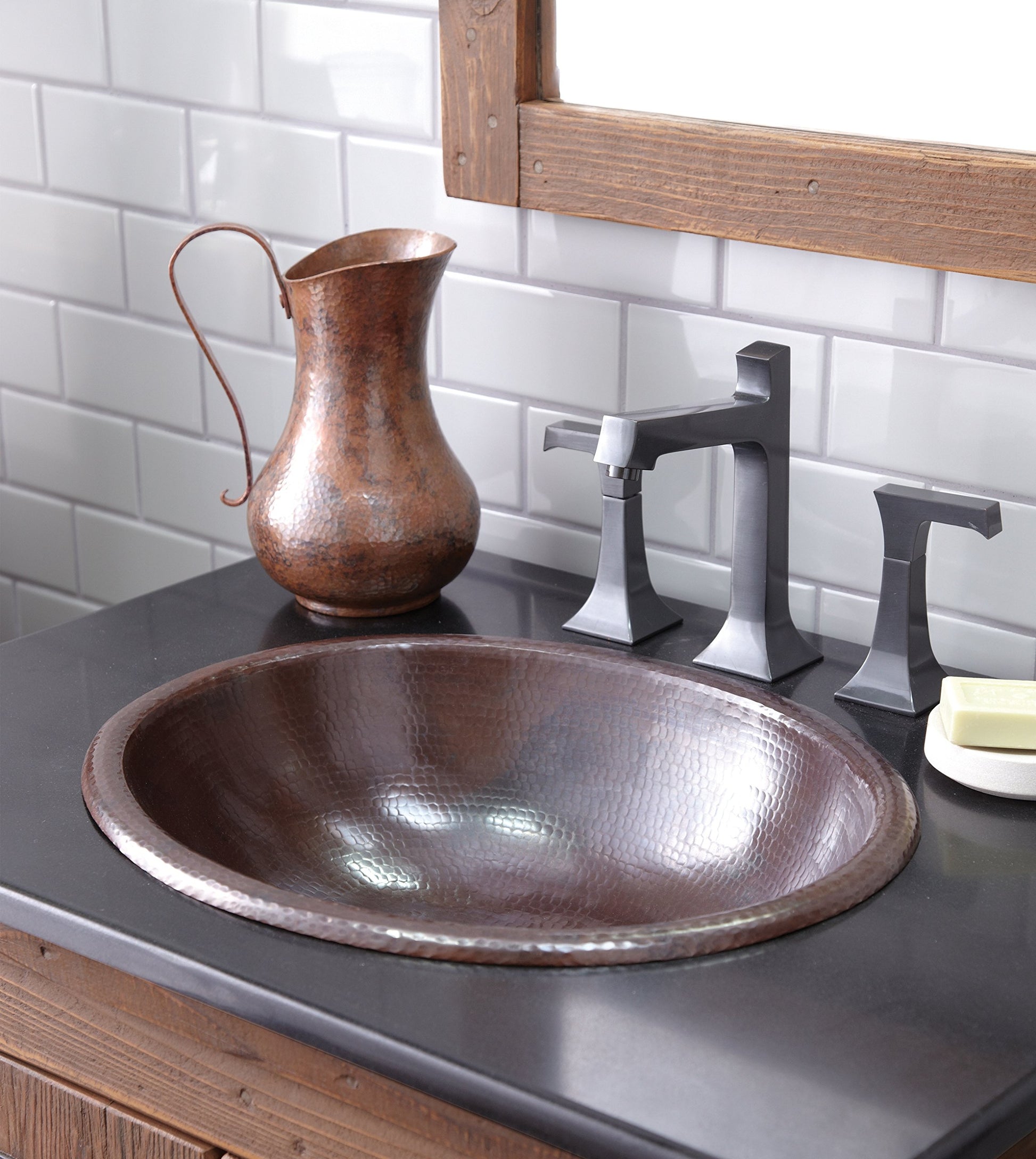Hammered copper oval bathroom sink with rolled rim on dark counter, paired with black faucet and copper pitcher.