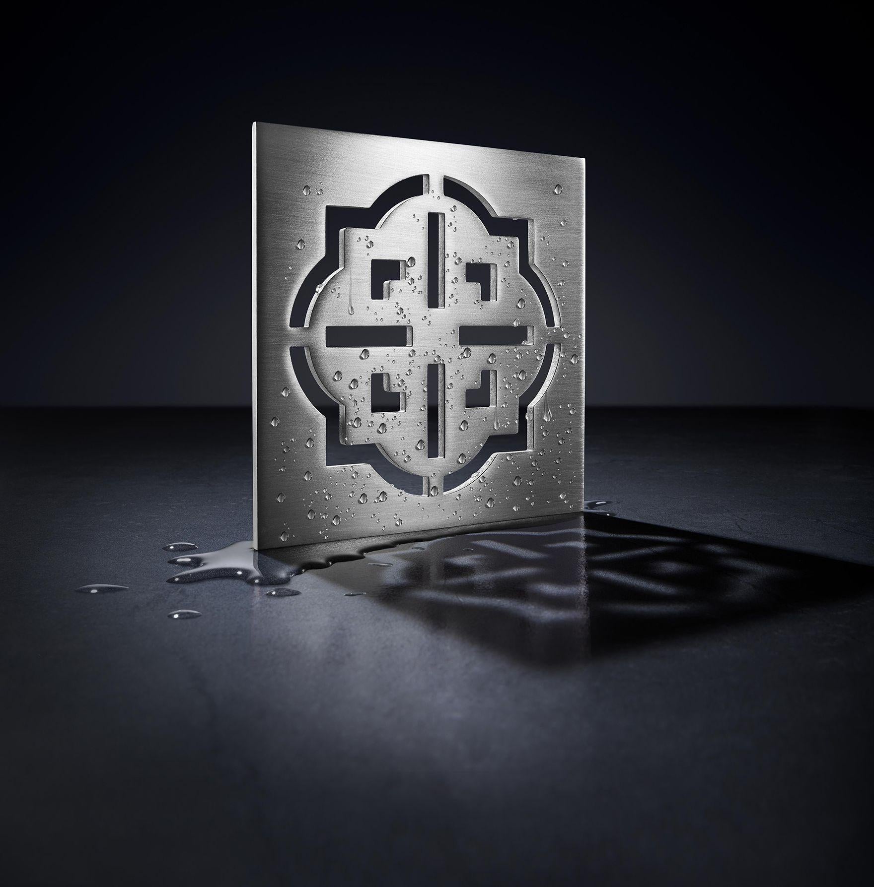 Burnished brass square drain trim grid with geometric quatrefoil pattern and mounting holes, displayed wet.