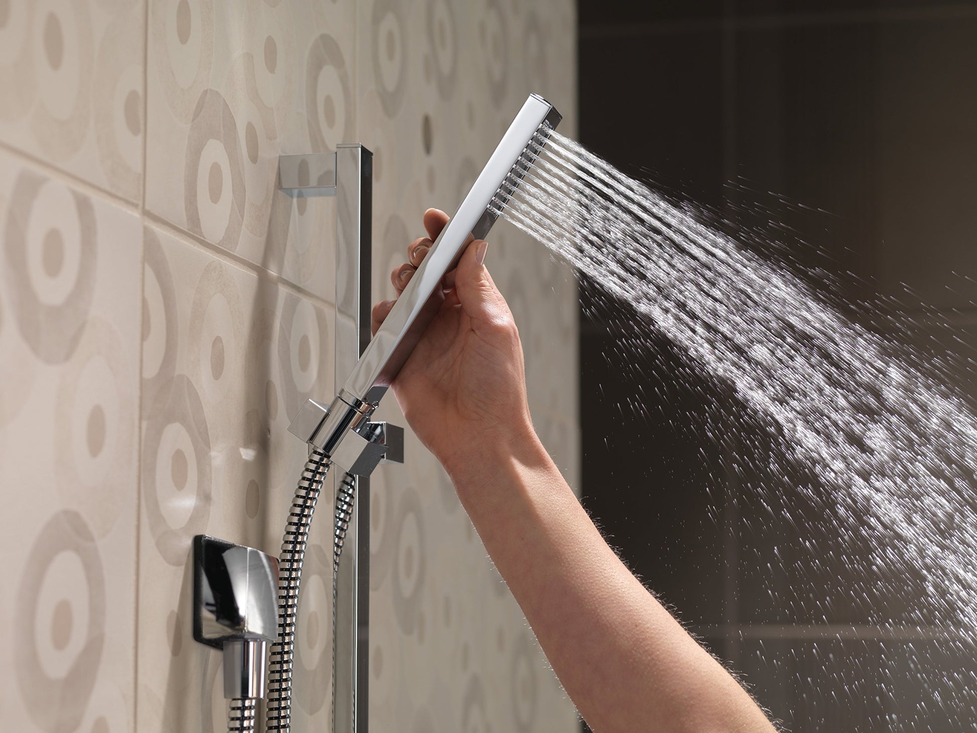 Chrome square wall elbow with hand shower head spraying water, mounted on tiled bathroom wall.