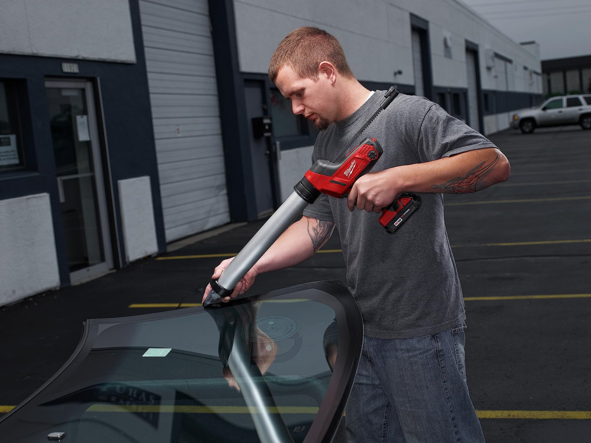 Man using red and silver caulking gun with aluminum barrel on car windshield in parking lot.