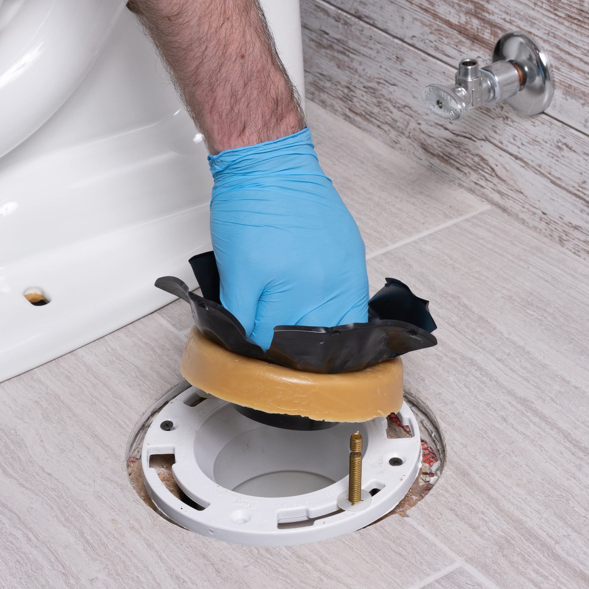 Hand in blue glove positioning tan wax ring with black flange onto white toilet base flange during installation.