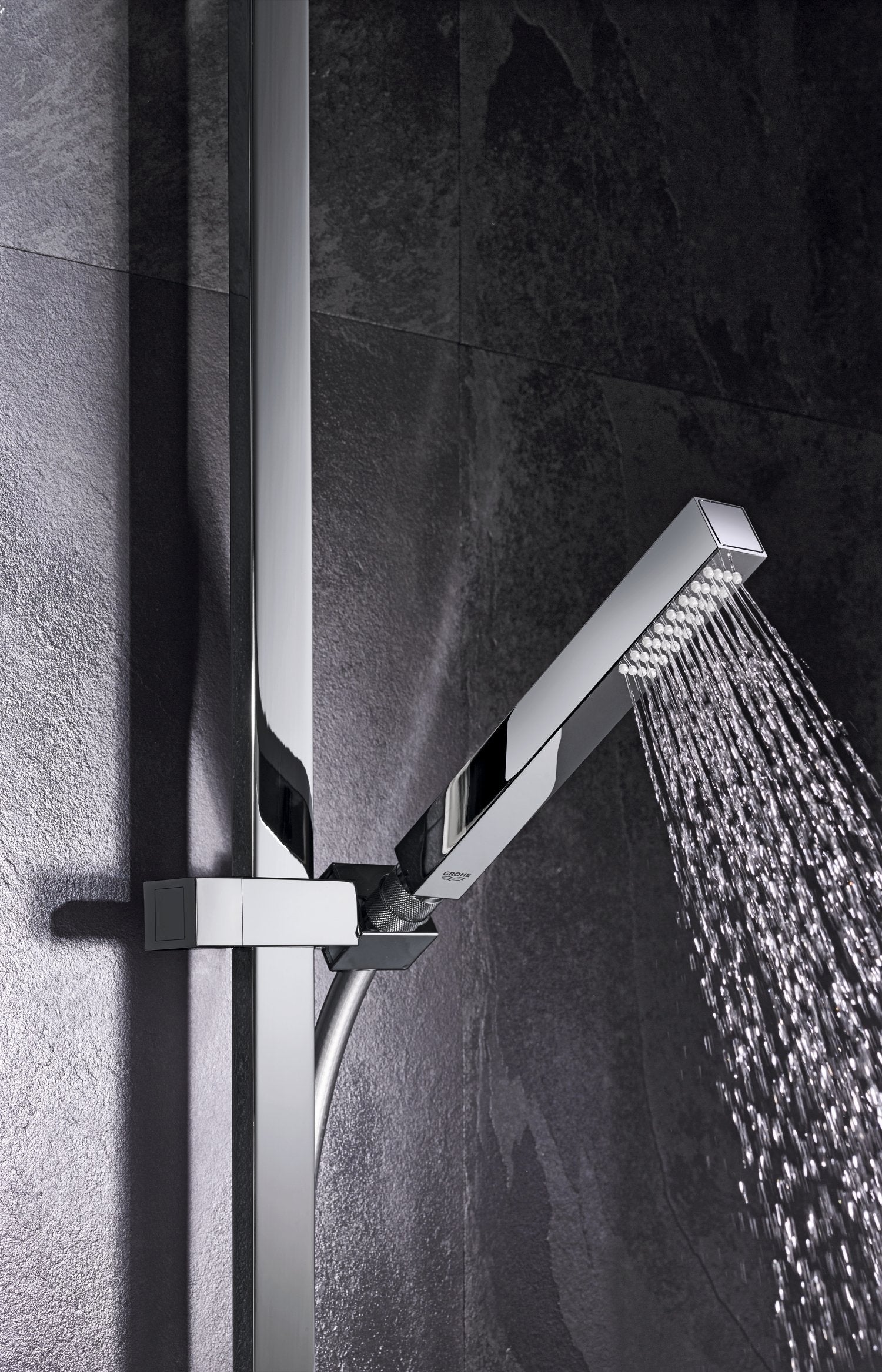 Starlight chrome shower bar with square head and handheld shower head spraying water against dark tile wall.