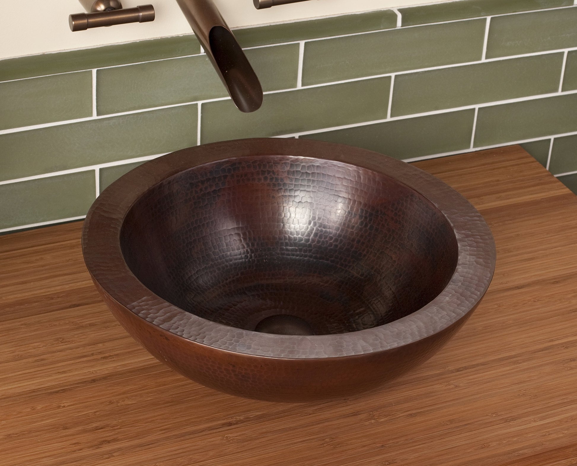 Hammered copper round vessel sink with antique dark finish on wooden countertop with green subway tile backsplash.