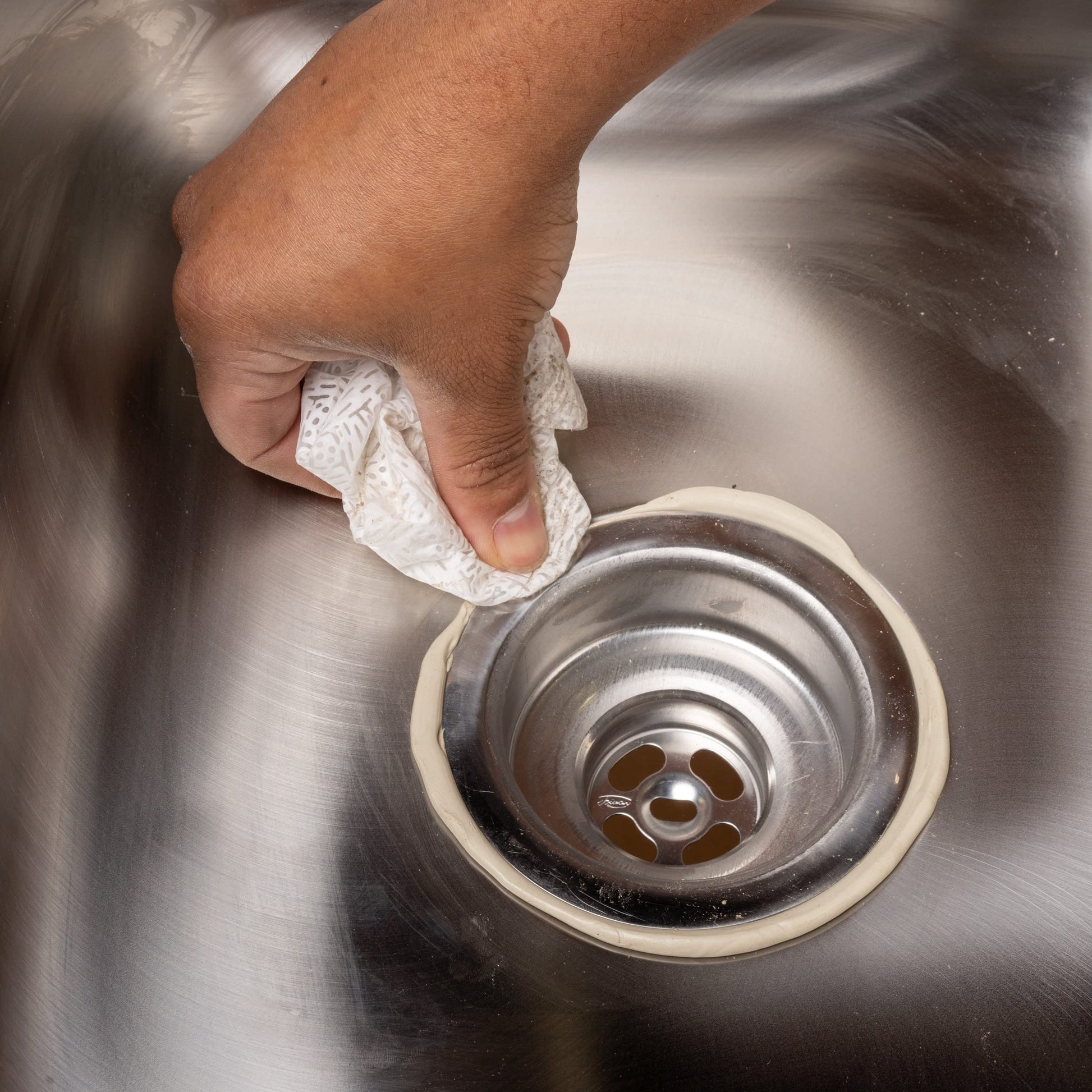 Tan-colored plumber's putty being applied around a sink drain opening with a hand holding the material.