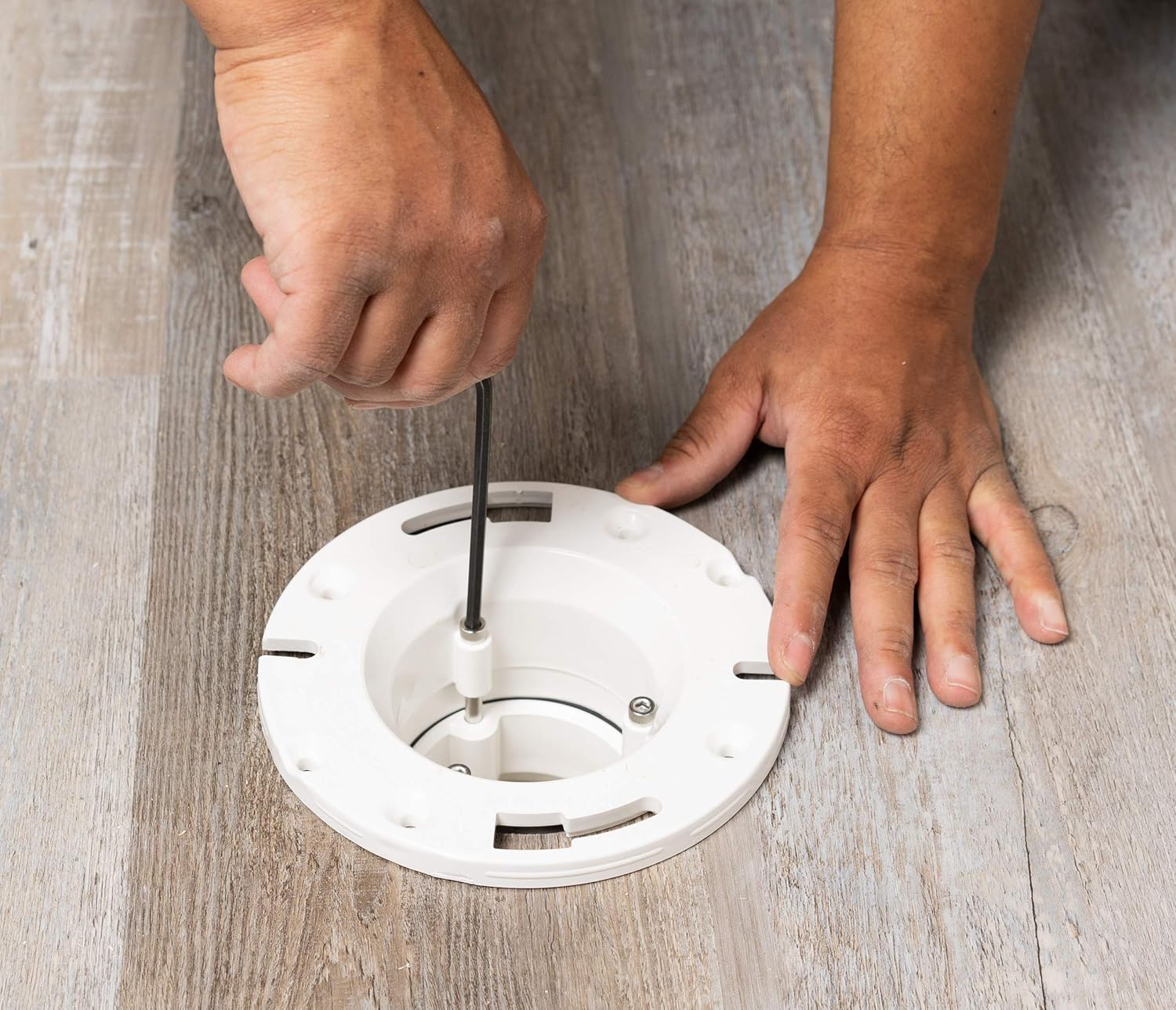 White PVC flange with center opening and mounting slots, shown being installed on wooden floor.