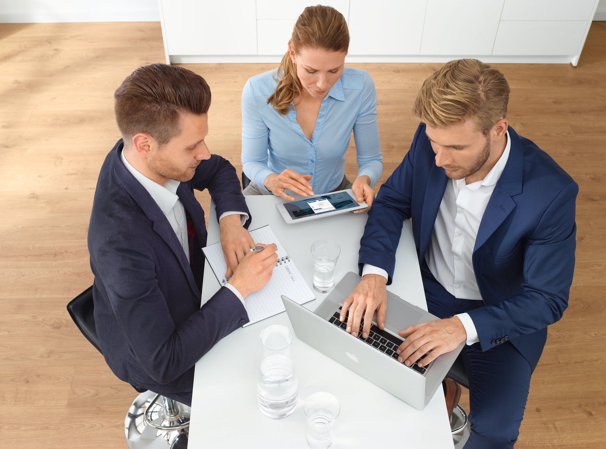 Six clear water glasses arranged on a white table with three business professionals around it.