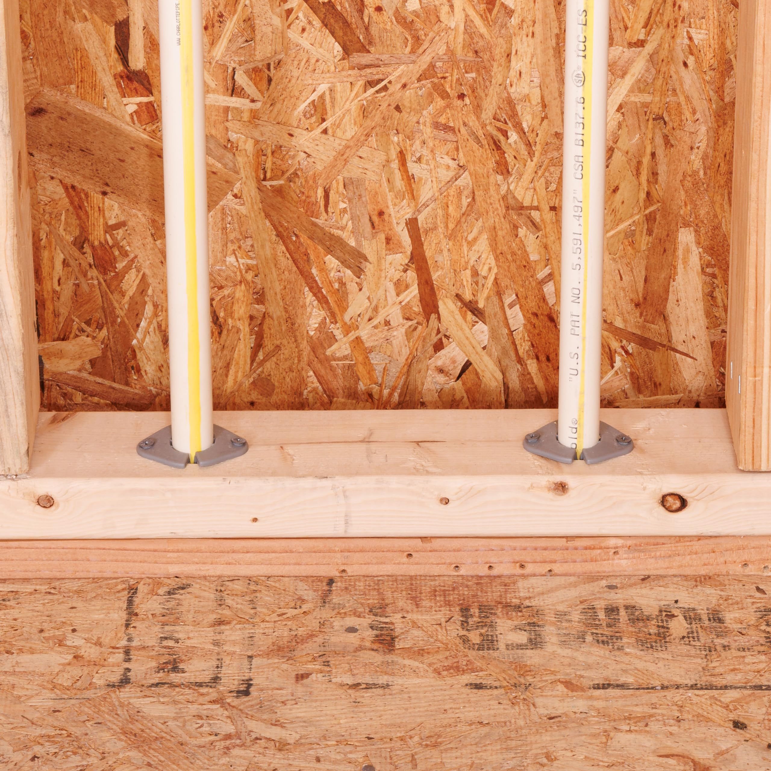 Two gray insulating pipe clamps securing yellow PEX pipes to wooden framing in construction.