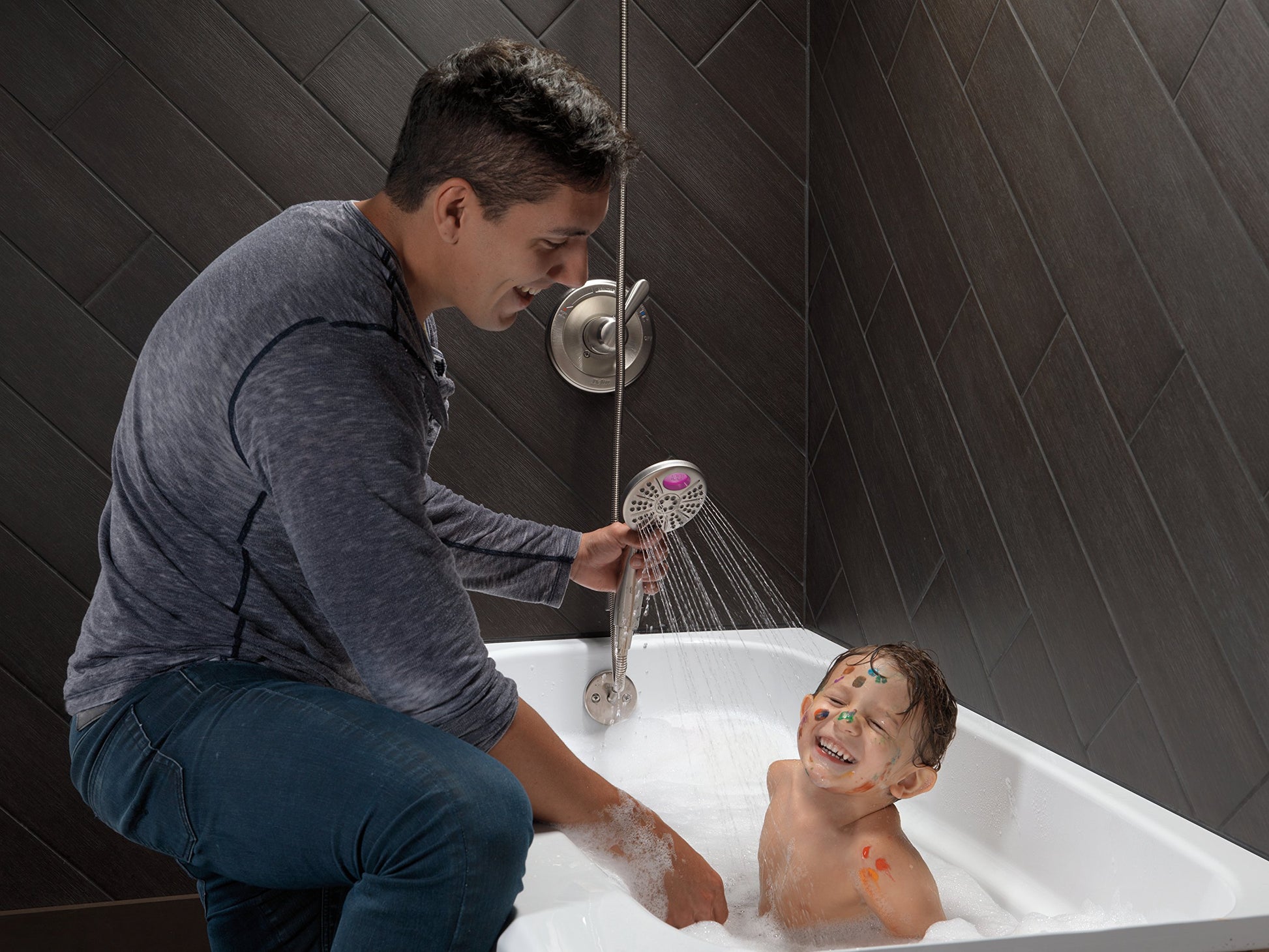 Stainless steel tub spout with pull-up diverter mounted on dark tile wall above white bathtub.