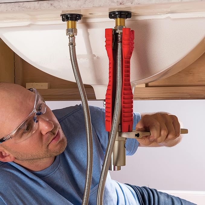 Plumber using red faucet removal tool under sink with supply lines and basin visible.