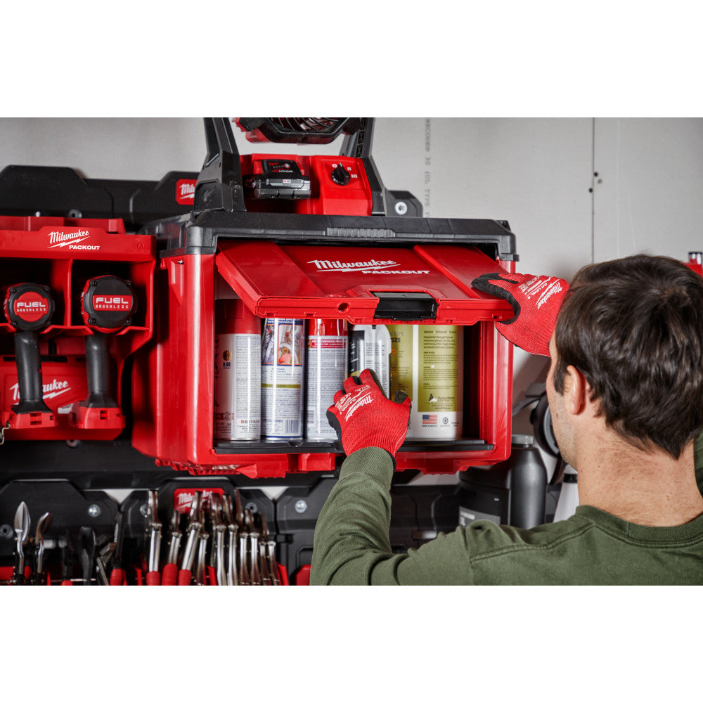 Red and black Milwaukee PACKOUT cabinet with open drawer displaying spray cans and supplies, person reaching inside wearing r