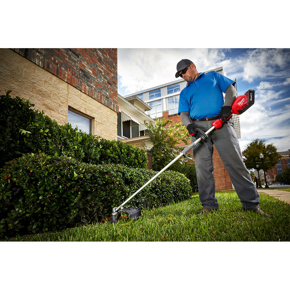 Man in blue shirt trimming grass and shrubs with a red and silver string trimmer near a brick house.