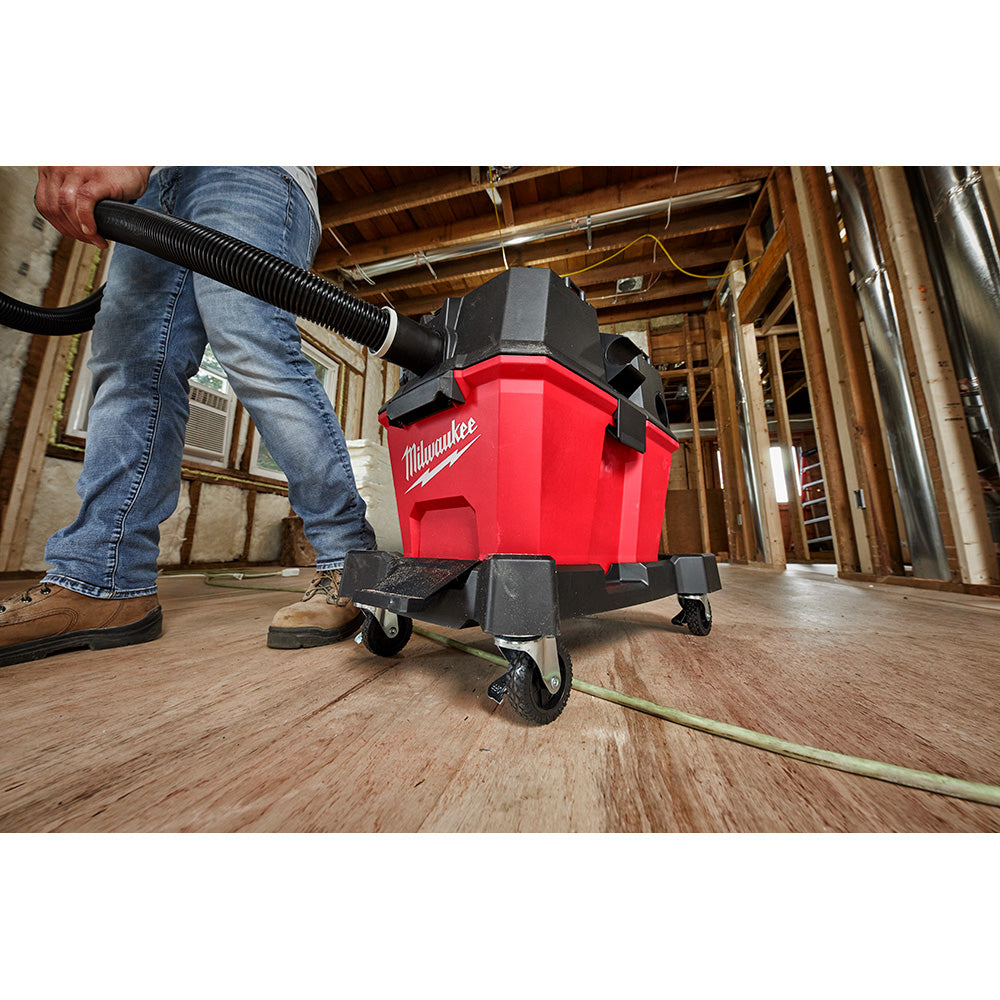 Red and black Milwaukee wet/dry vacuum cart with hose being held by a person in jeans on a wooden floor.