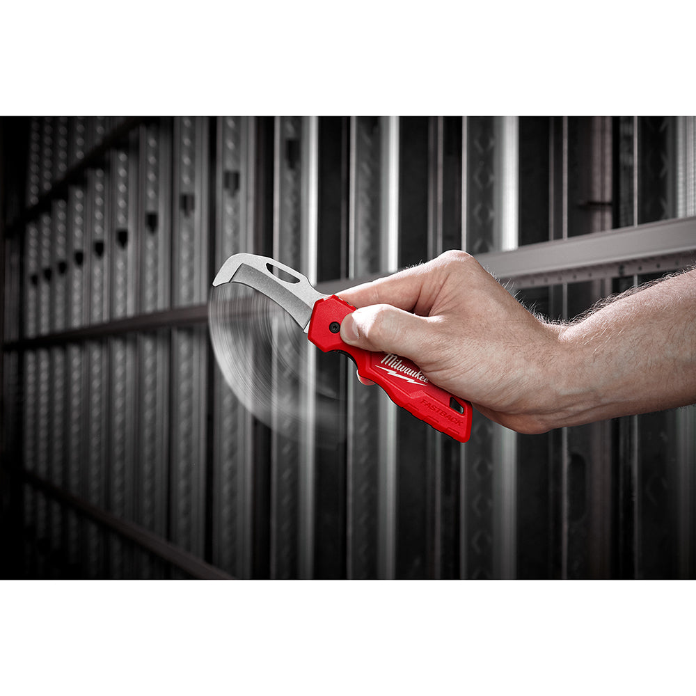 Red and silver folding knife with curved hawkbill blade held in a hand against industrial background.