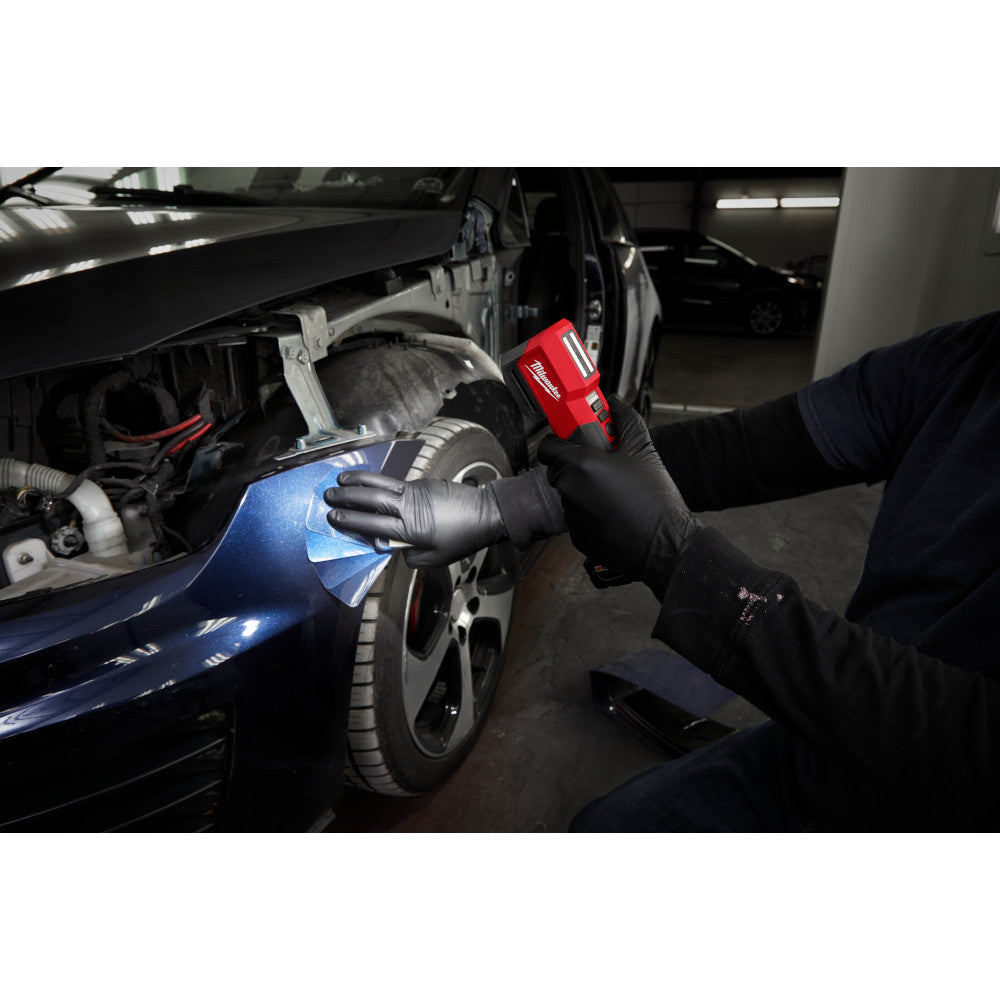 Red Milwaukee M12 paint light held by gloved hand shining on blue car body panel in garage setting.