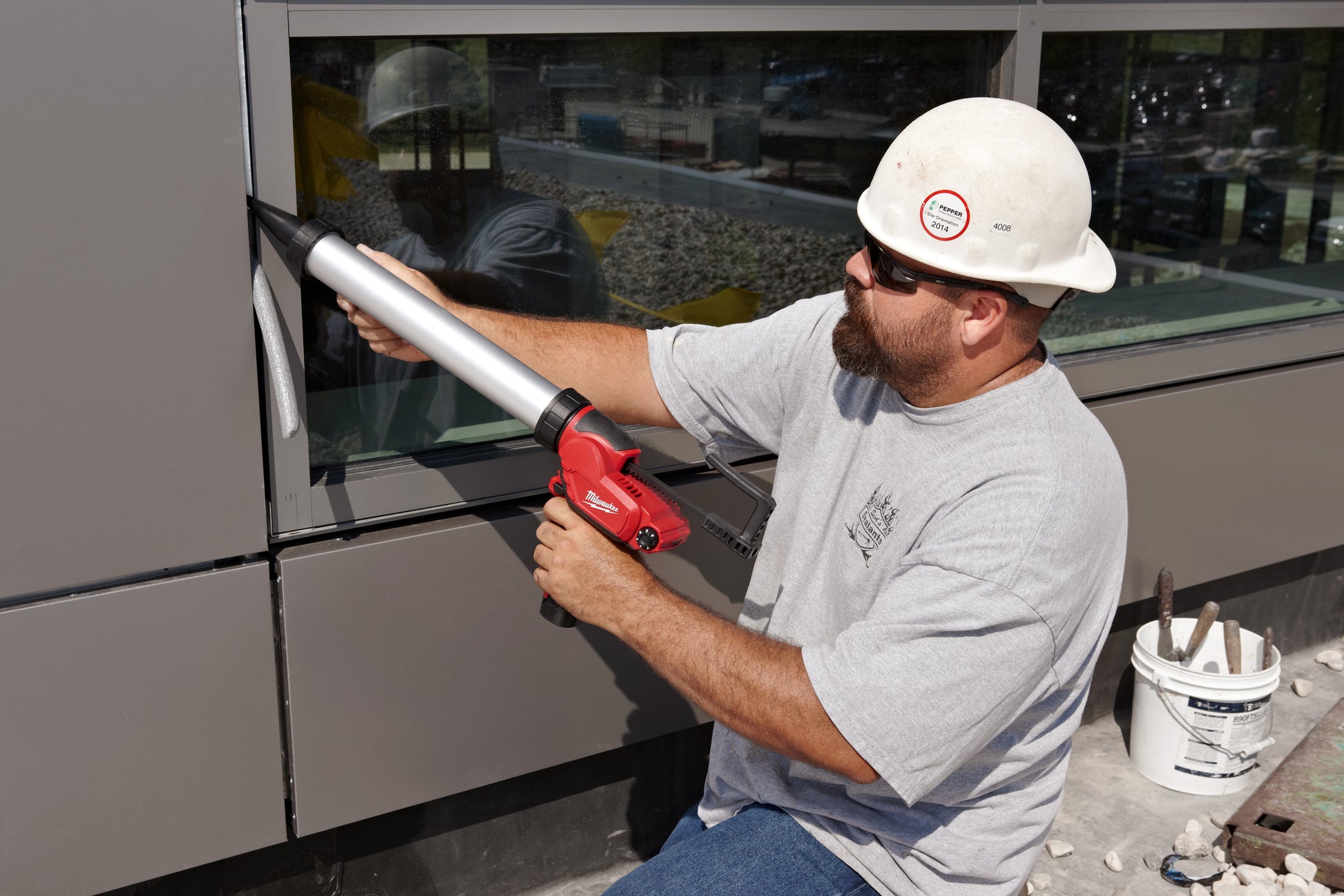 Worker holding red caulking gun with aluminum barrel while applying sealant to exterior panel.