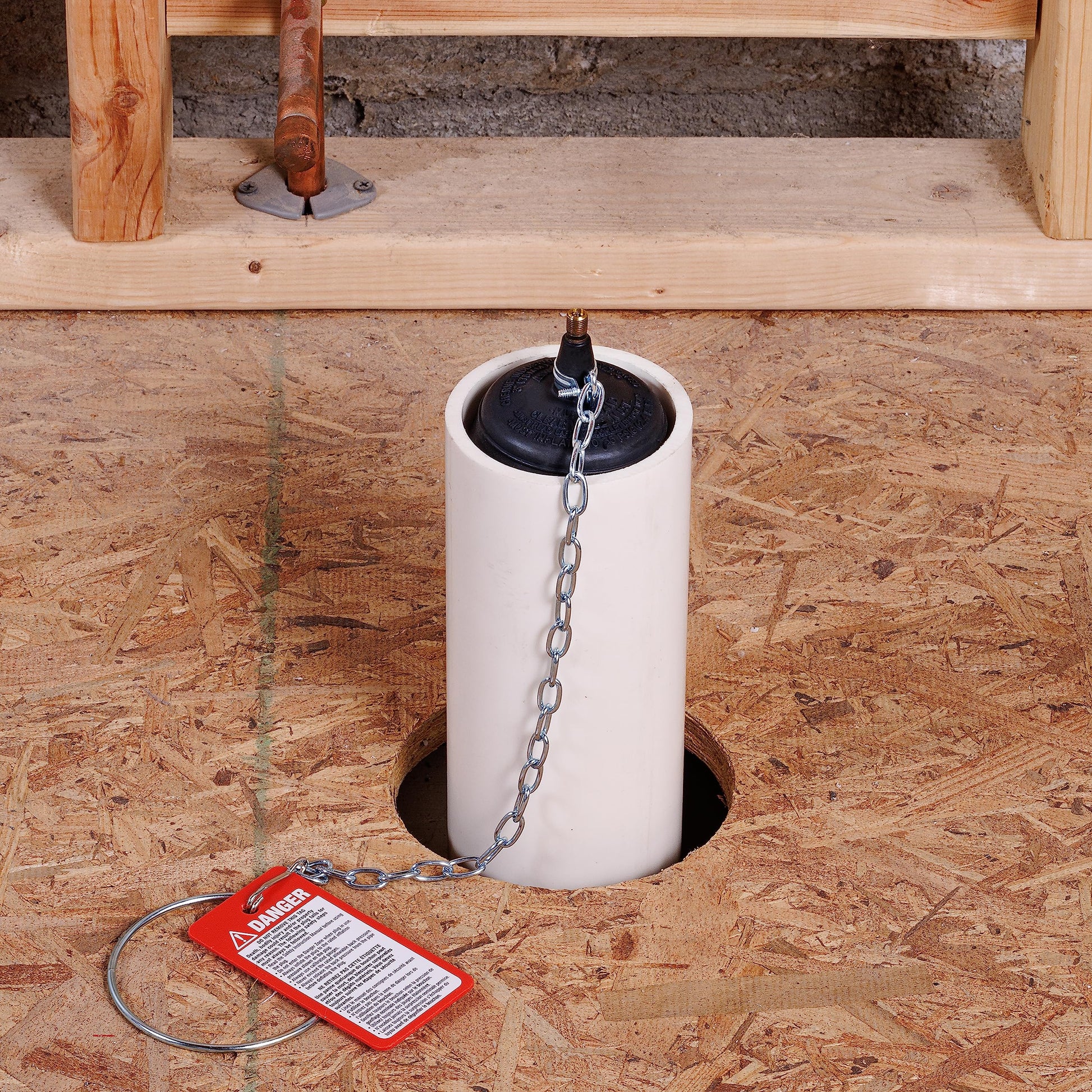 White cylindrical test-ball plug with black cap and chain attached, displayed in concrete opening with red warning tag.