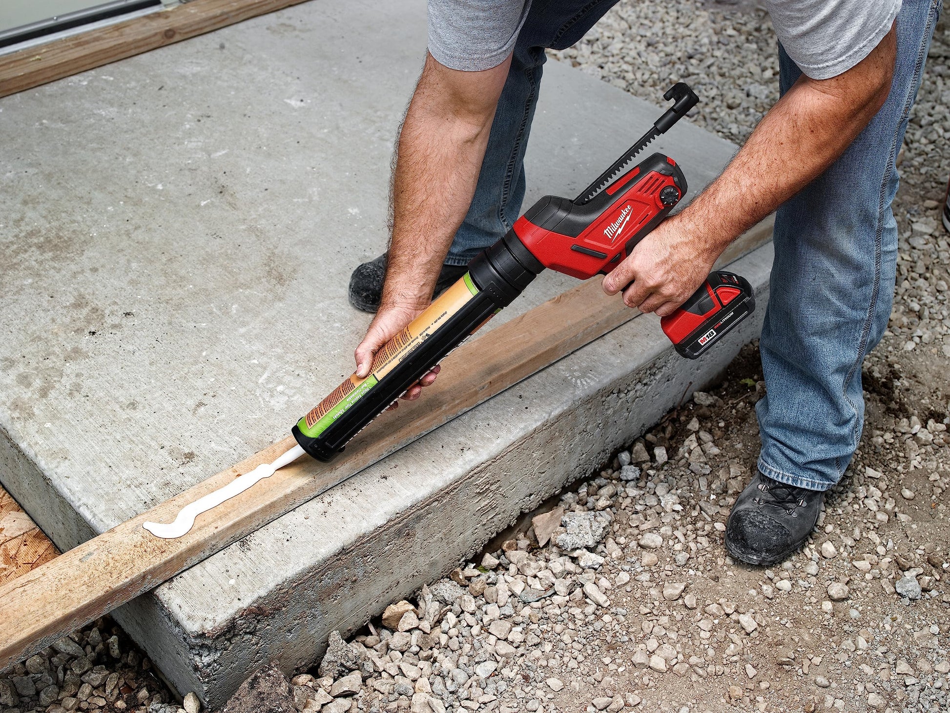 Person using red and black Milwaukee caulking gun with aluminum barrel on wooden trim joint.
