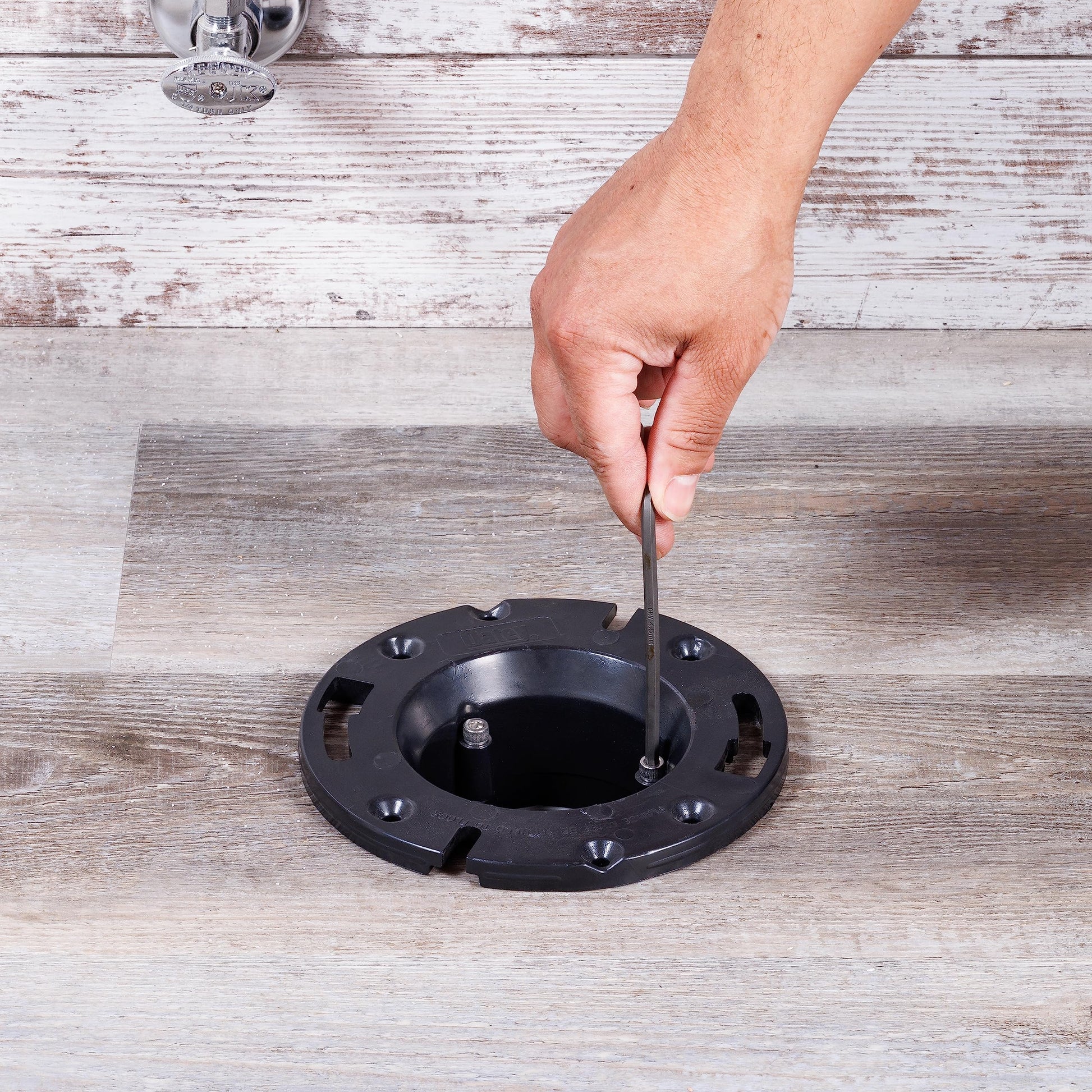 Black ABS closet flange with mounting holes and bolt being inserted by hand on wooden floor.