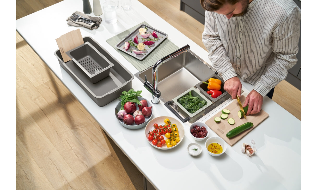 Stainless steel sink system with multiple compartments, strainers, and accessories arranged on white counter with fresh veget