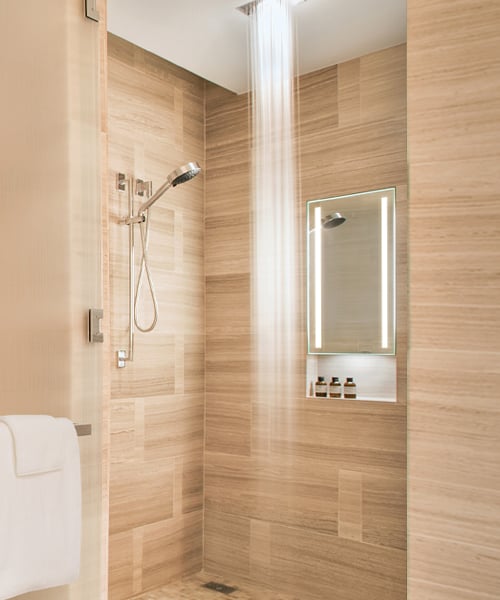Illuminated rectangular bathroom mirror with white LED border mounted on wood-tile wall above shelf with toiletries.