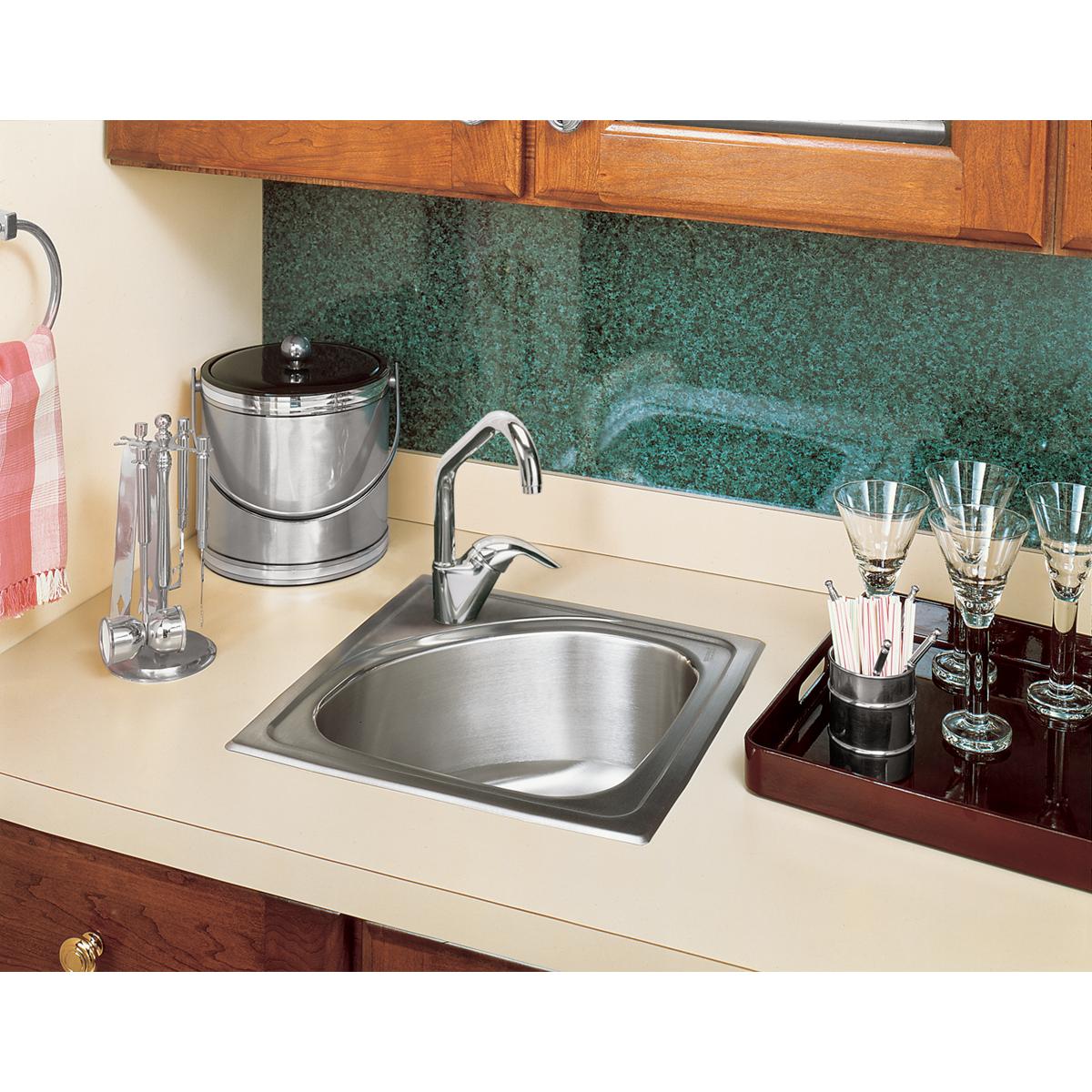 Stainless steel square drop-in bar sink with single faucet hole, installed in beige countertop next to green backsplash.