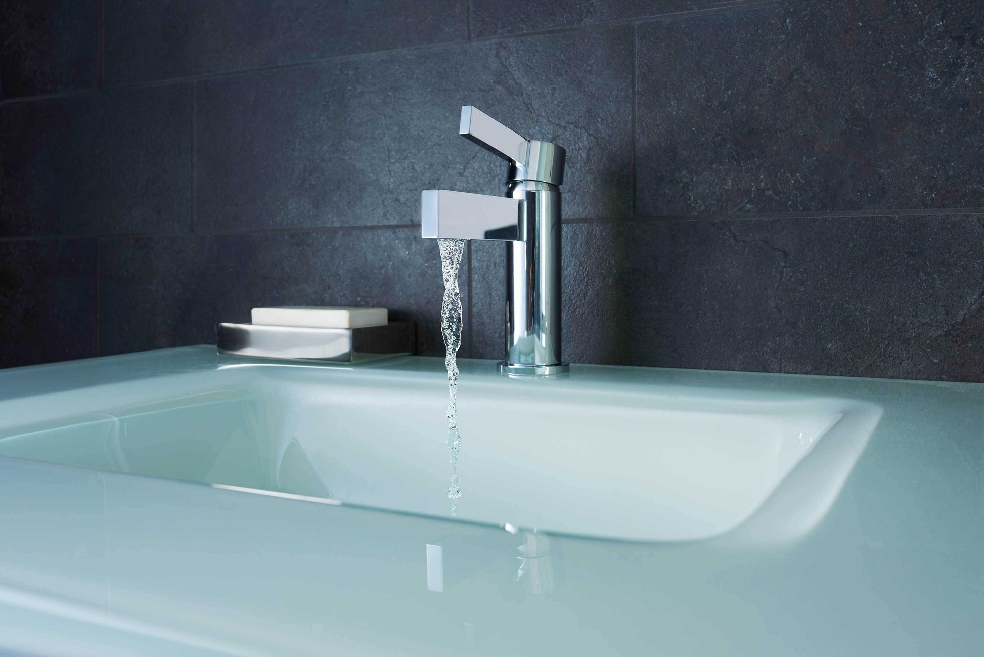 Polished chrome single-hole lavatory faucet with angular handle mounted on glass countertop above white sink.