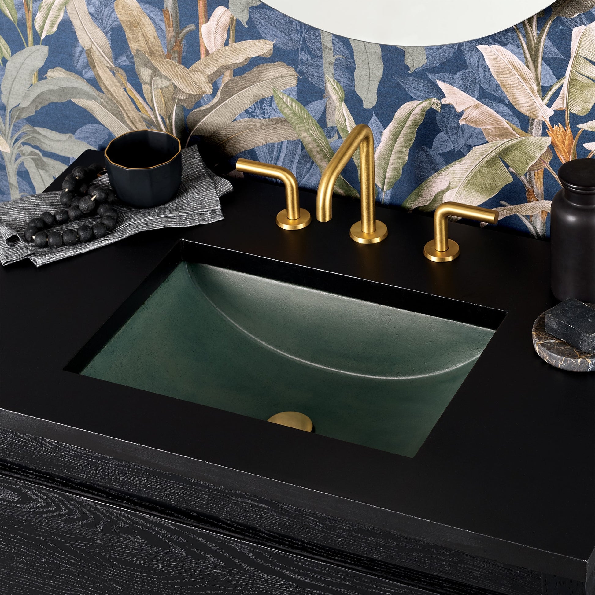 Green undermount sink with brass faucet and handles on black countertop, blue botanical wallpaper backdrop.
