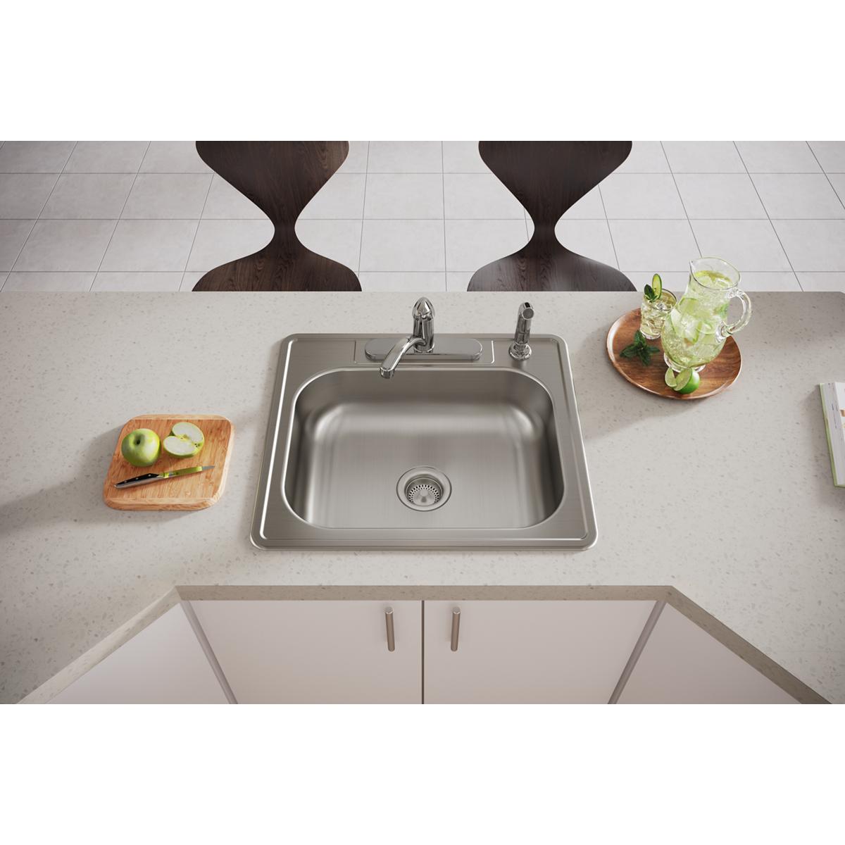 Stainless steel single bowl drop-in sink with four holes mounted in light countertop, flanked by cutting board and beverage g