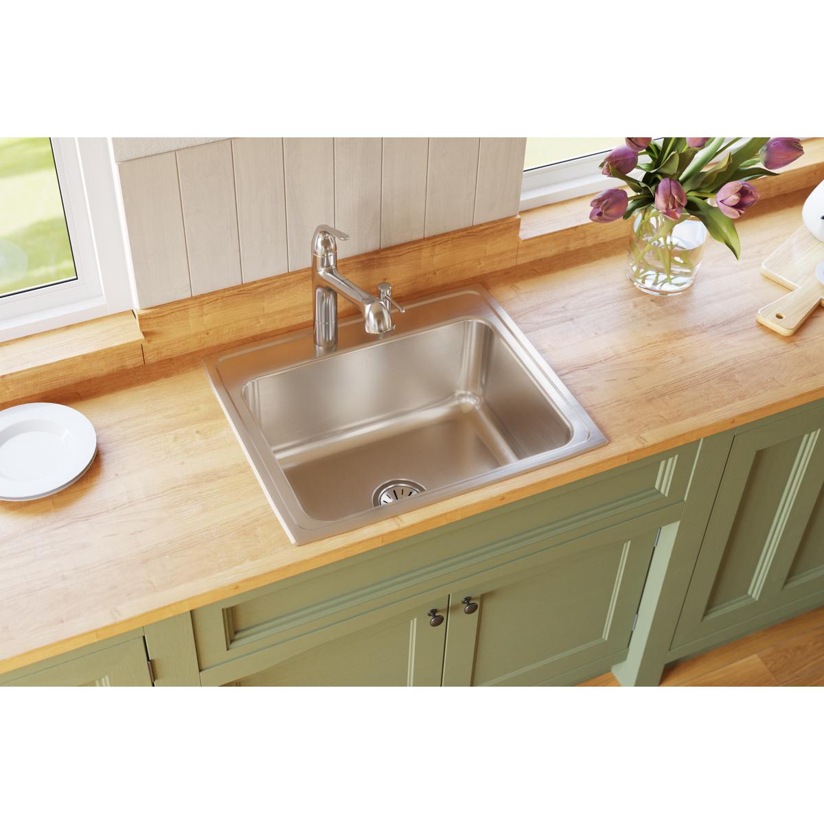 Stainless steel single bowl drop-in sink with two faucet holes installed in wooden countertop above green cabinetry.