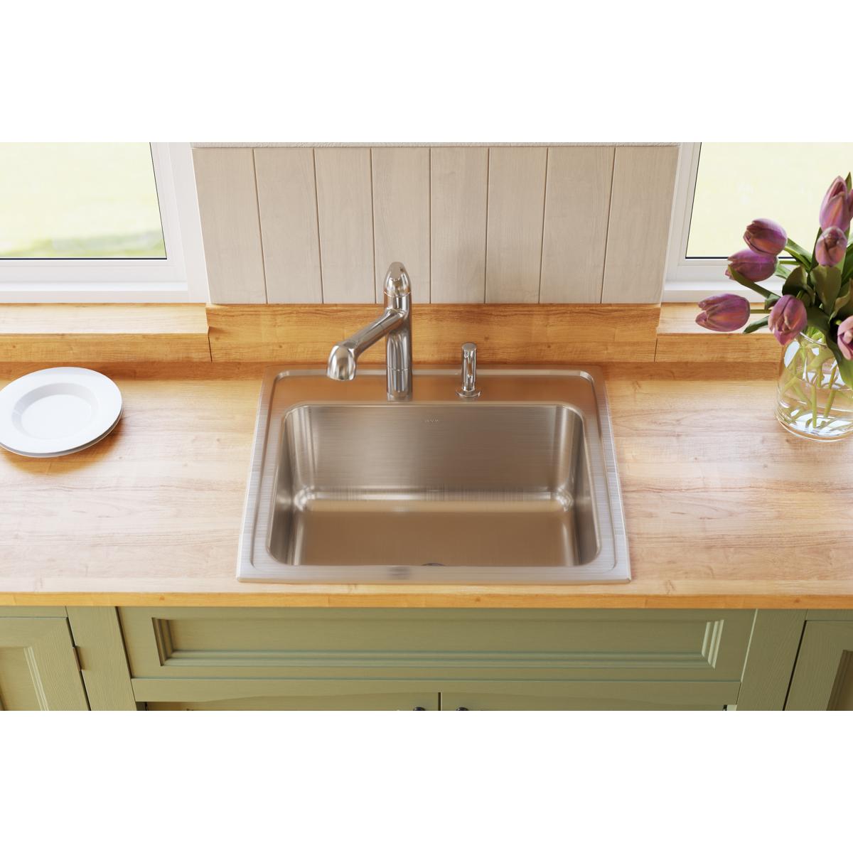 Stainless steel drop-in sink with single bowl and two faucet holes installed in wooden countertop with chrome faucet.