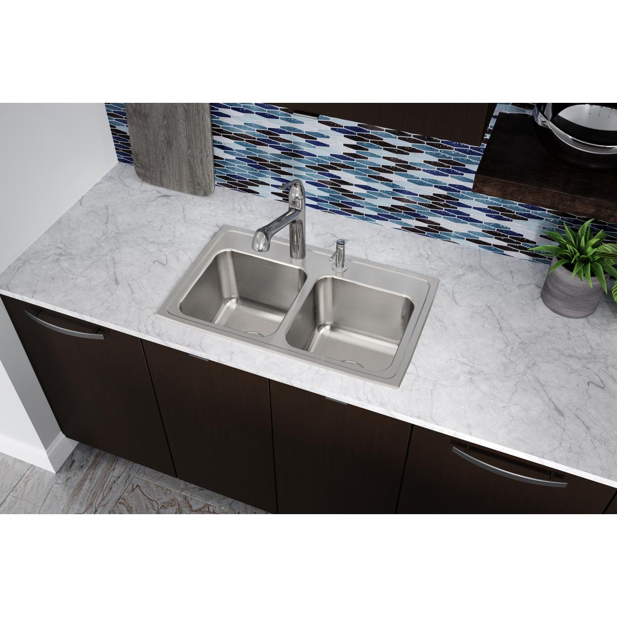 Stainless steel double bowl drop-in sink with five holes installed in white marble countertop above dark cabinetry.