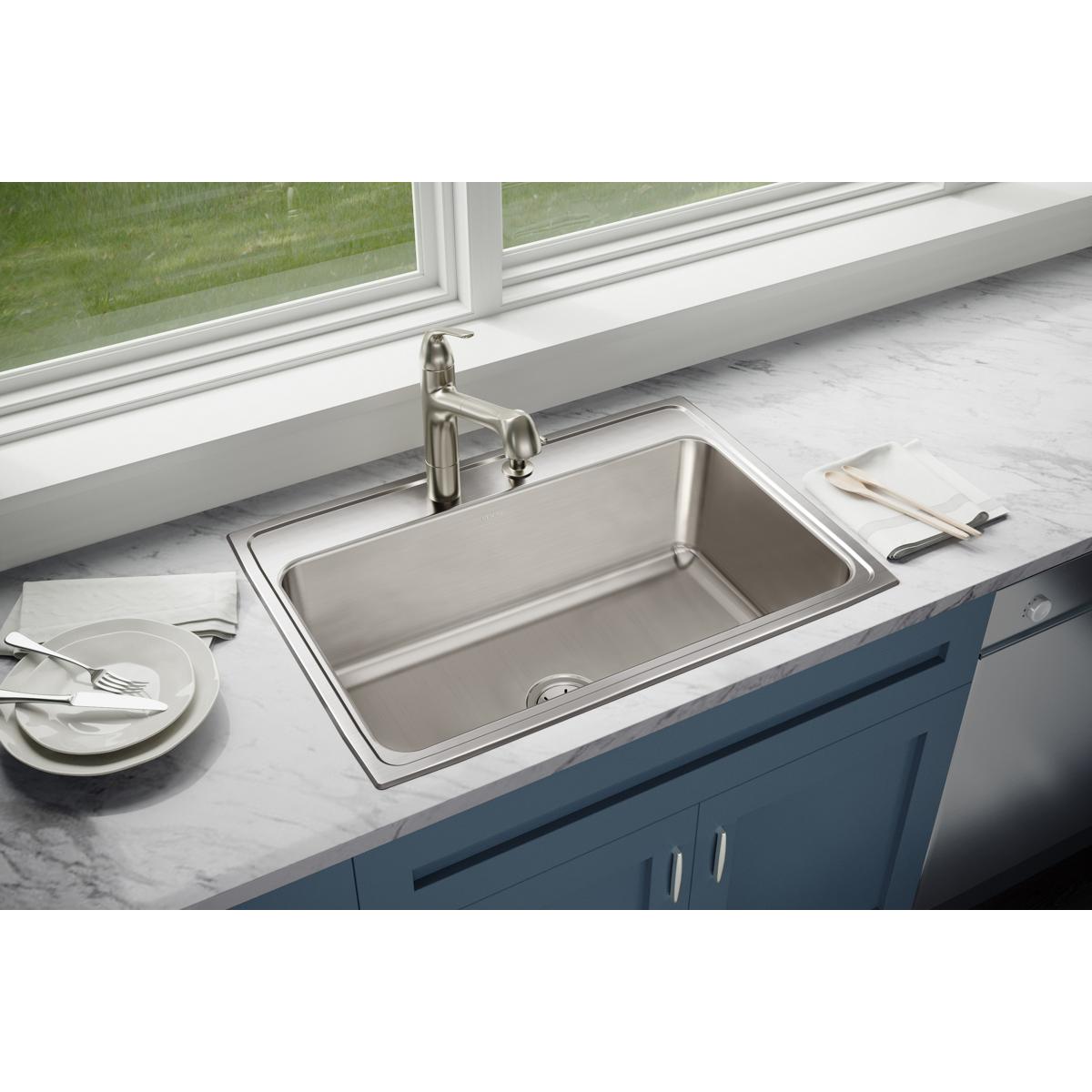 Stainless steel single bowl drop-in sink with faucet installed in marble countertop next to window.