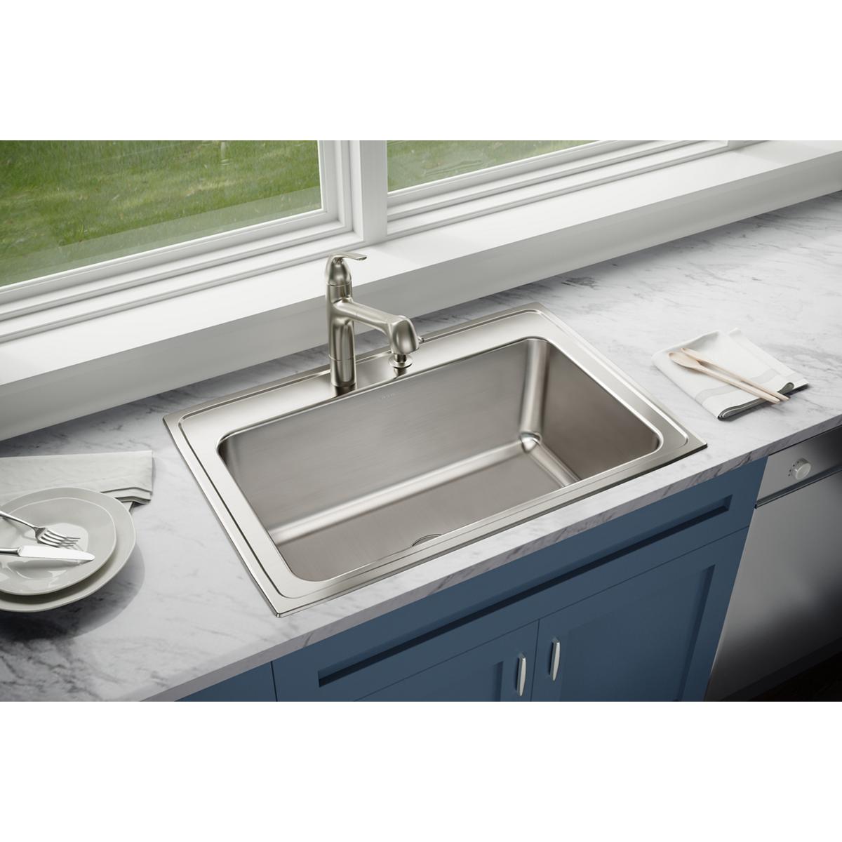 Stainless steel drop-in kitchen sink with single bowl and two faucet holes, installed in white countertop.